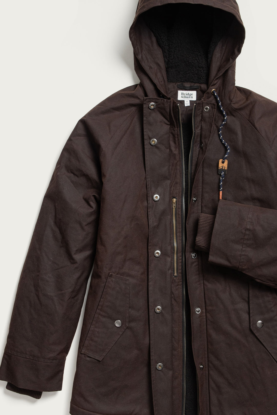 The Sequoia Parka by Bridge & Burn features umber waxed canvas, snap buttons, front pockets, ribbed cuffs, and a drawstring with a wooden toggle, shown flat against a light background.