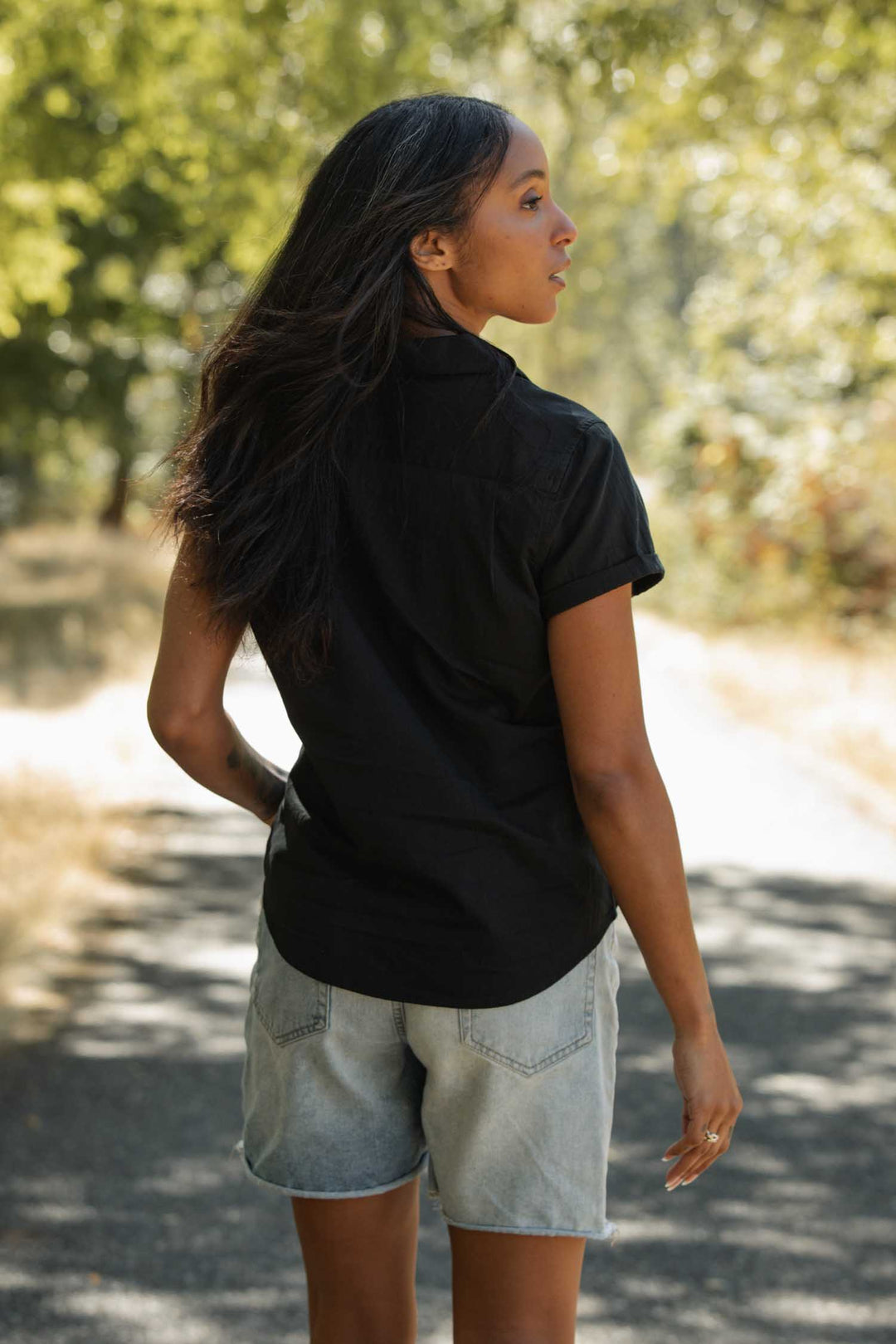 A woman with long dark hair wears the Bridge & Burn Innes Shirt in black and light denim shorts, standing on a sunlit path with lush greenery in the background, looking to her left.