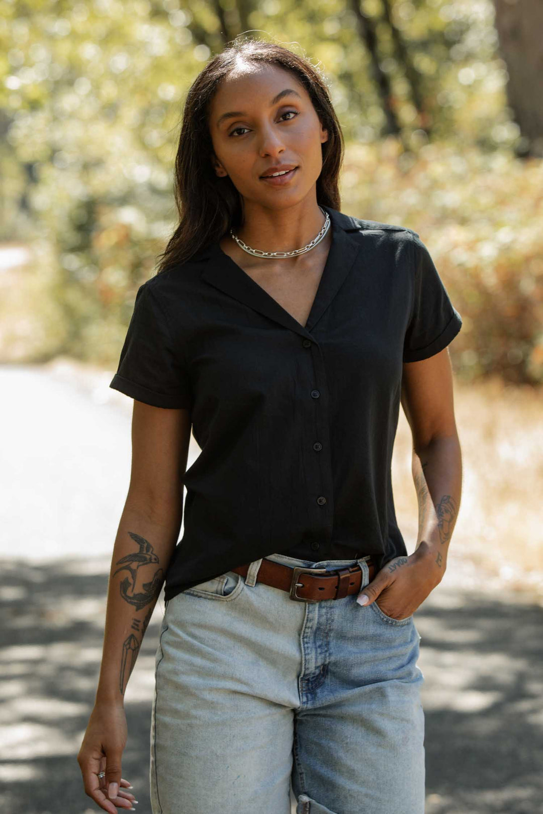 A woman with long dark hair wears the Bridge & Burn Innes Shirt in black, paired with light blue jeans and a chain necklace, as she stands outdoors on a sunlit path surrounded by trees and greenery.