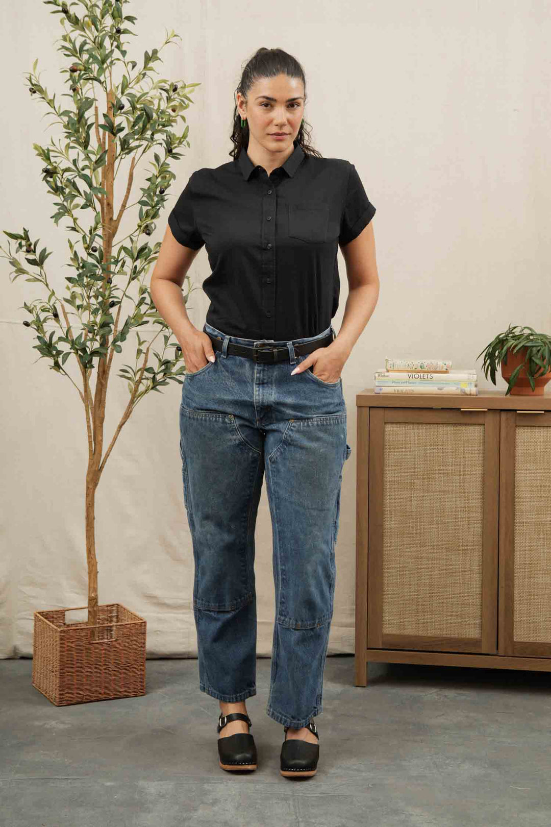 A woman stands indoors with her hands in her pockets, wearing the Bridge & Burn Bea Shirt in black, blue jeans, and black sandals. A tall potted plant and a cabinet with books and a small plant are nearby.
