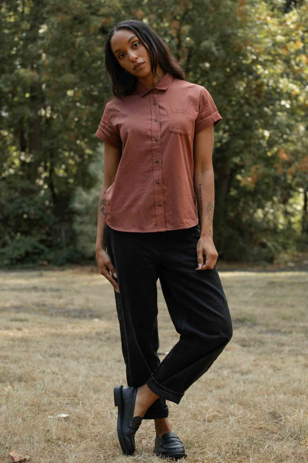 A woman with long hair and forearm tattoos stands on dry grass in front of green trees, wearing the Bridge & Burn Bea Shirt in Copper, paired with black pants and black shoes.