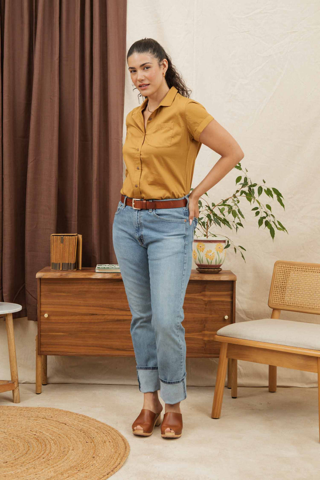 A woman wears the Bridge & Burn Bea Shirt in Gold, paired with light blue jeans and brown heels, standing in a modern room featuring wooden furniture, a plant, and neutral-colored curtains.