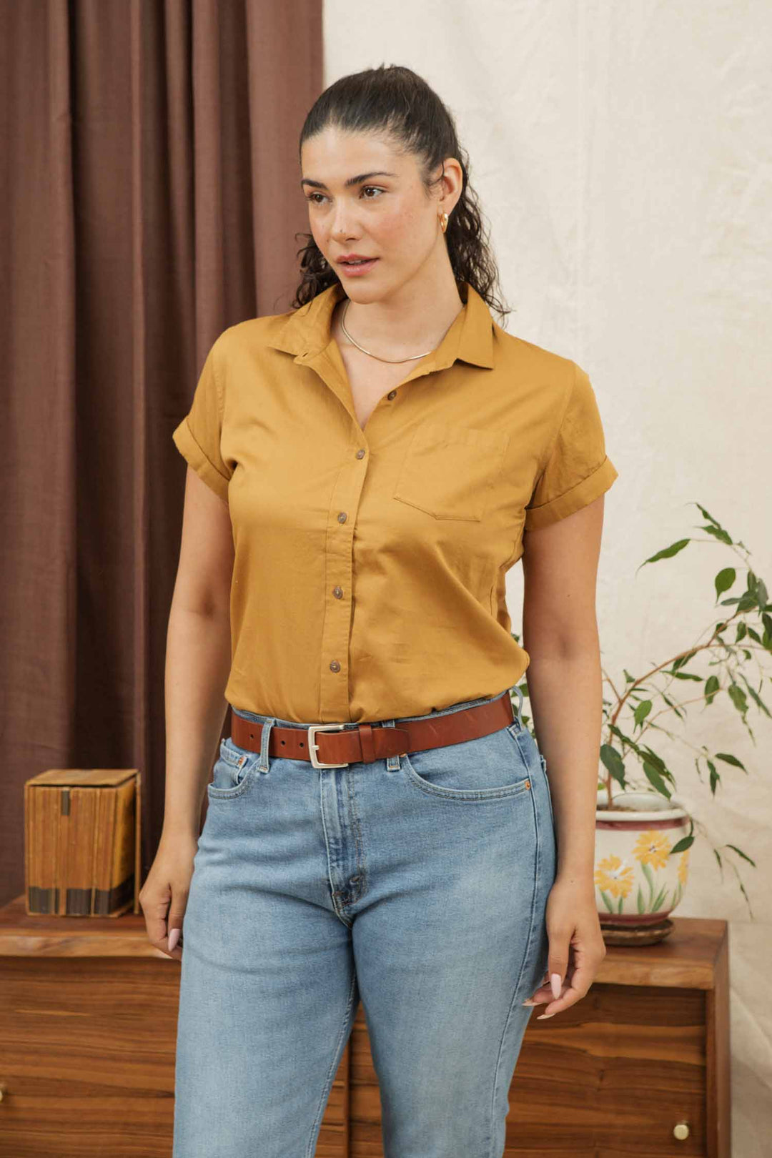 A woman with dark, curly hair tied back wears the Bridge & Burn Bea Shirt in Gold, a women’s short-sleeve button-down made from organic cotton, paired with light blue jeans. She stands indoors near a wooden table with decor and a potted plant.