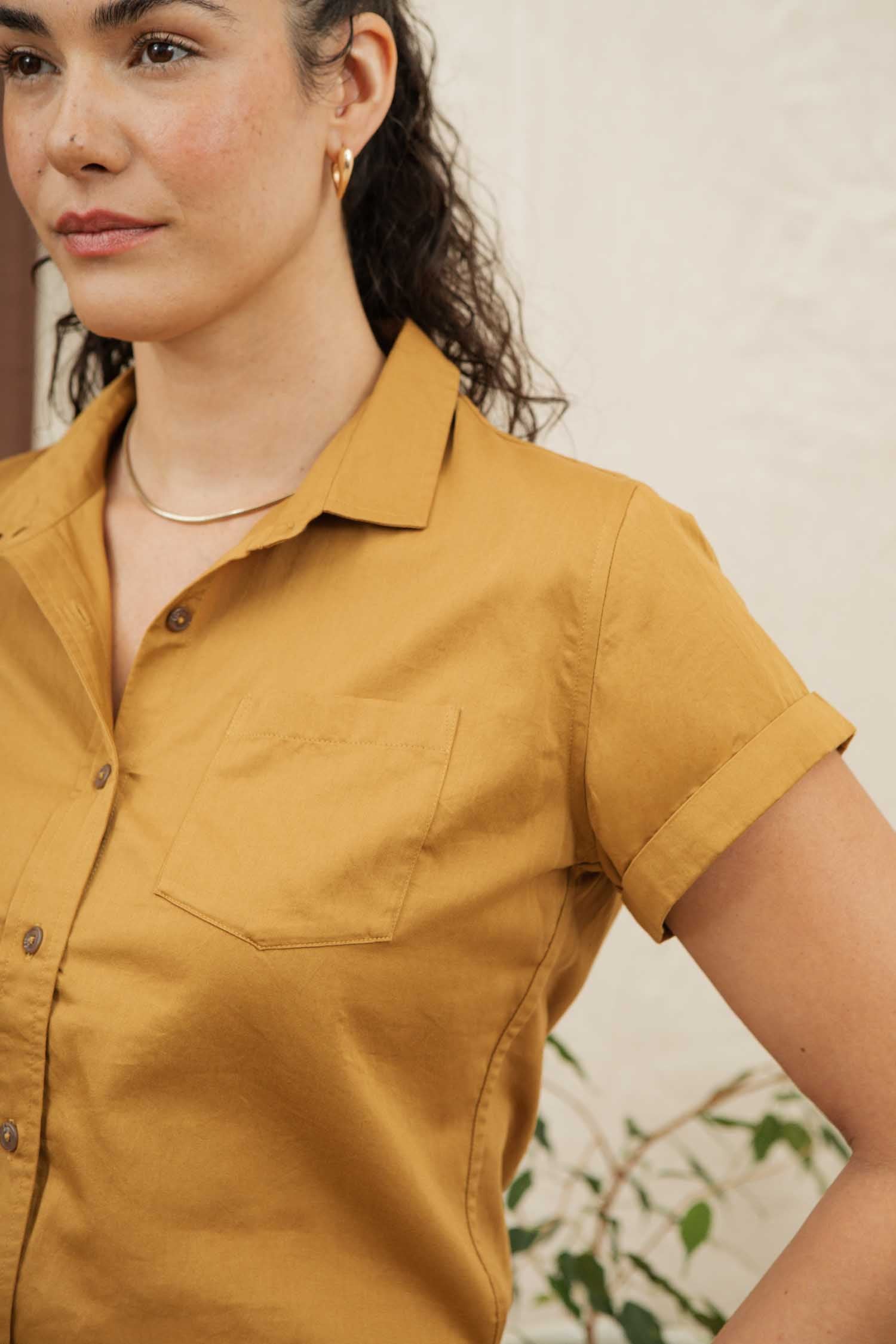 A woman with curly hair pulled back wears the Bridge & Burn Bea Shirt in Gold—an organic cotton short-sleeve button down—paired with a gold necklace and hoop earring, standing against a light background and turned partially to the side.