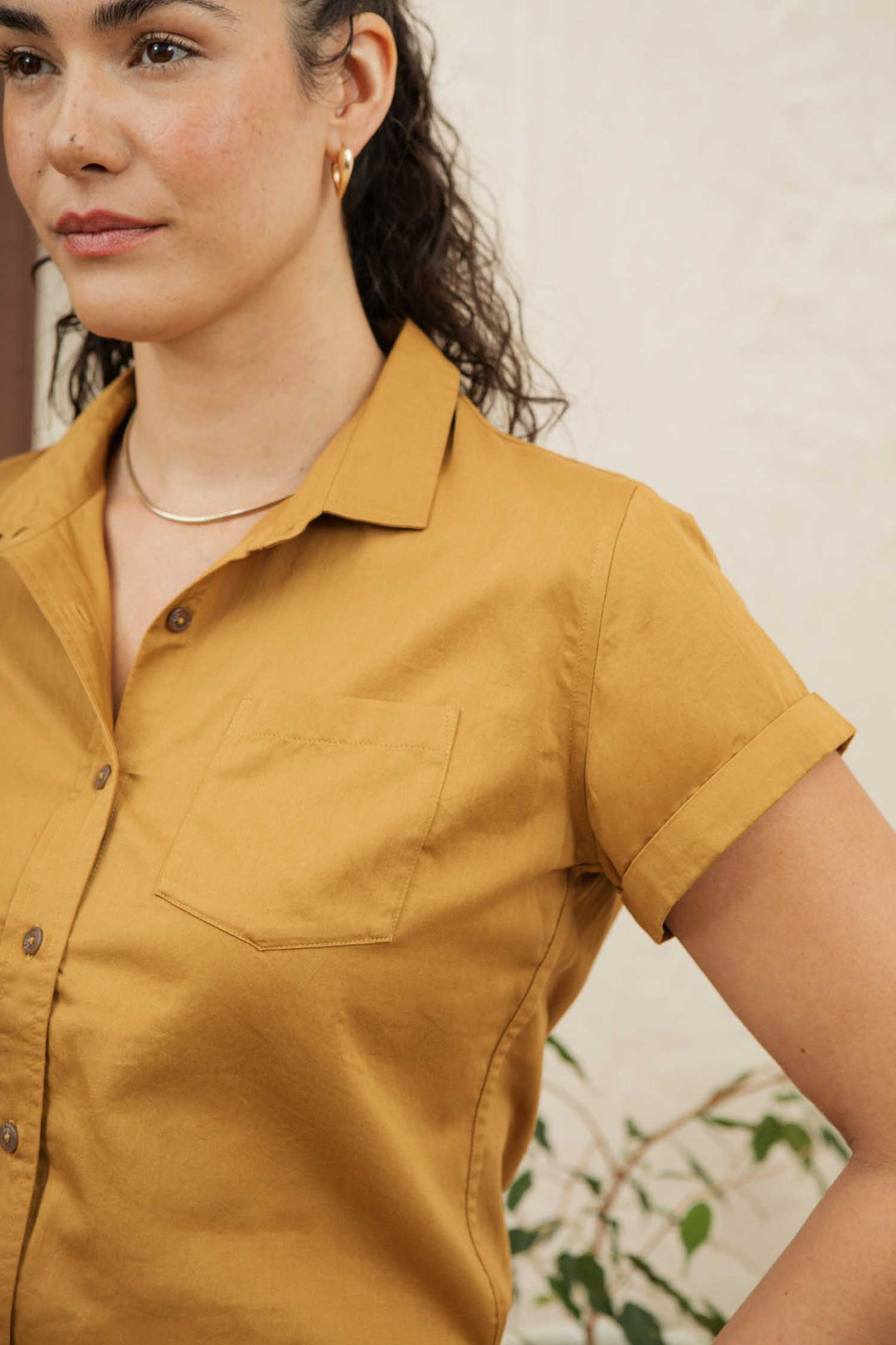 A woman with curly hair pulled back wears the Bridge & Burn Bea Shirt in Gold—an organic cotton short-sleeve button down—paired with a gold necklace and hoop earring, standing against a light background and turned partially to the side.