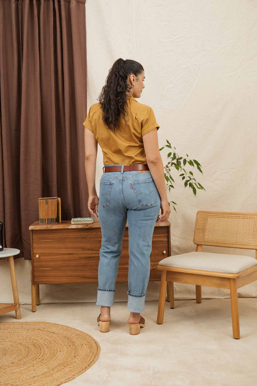 A woman with long, dark hair in a ponytail stands with her back to the camera, wearing the Bridge & Burn Bea Shirt in Gold, light blue cuffed jeans, and tan heels in a cozy room with wooden furniture and a potted plant.