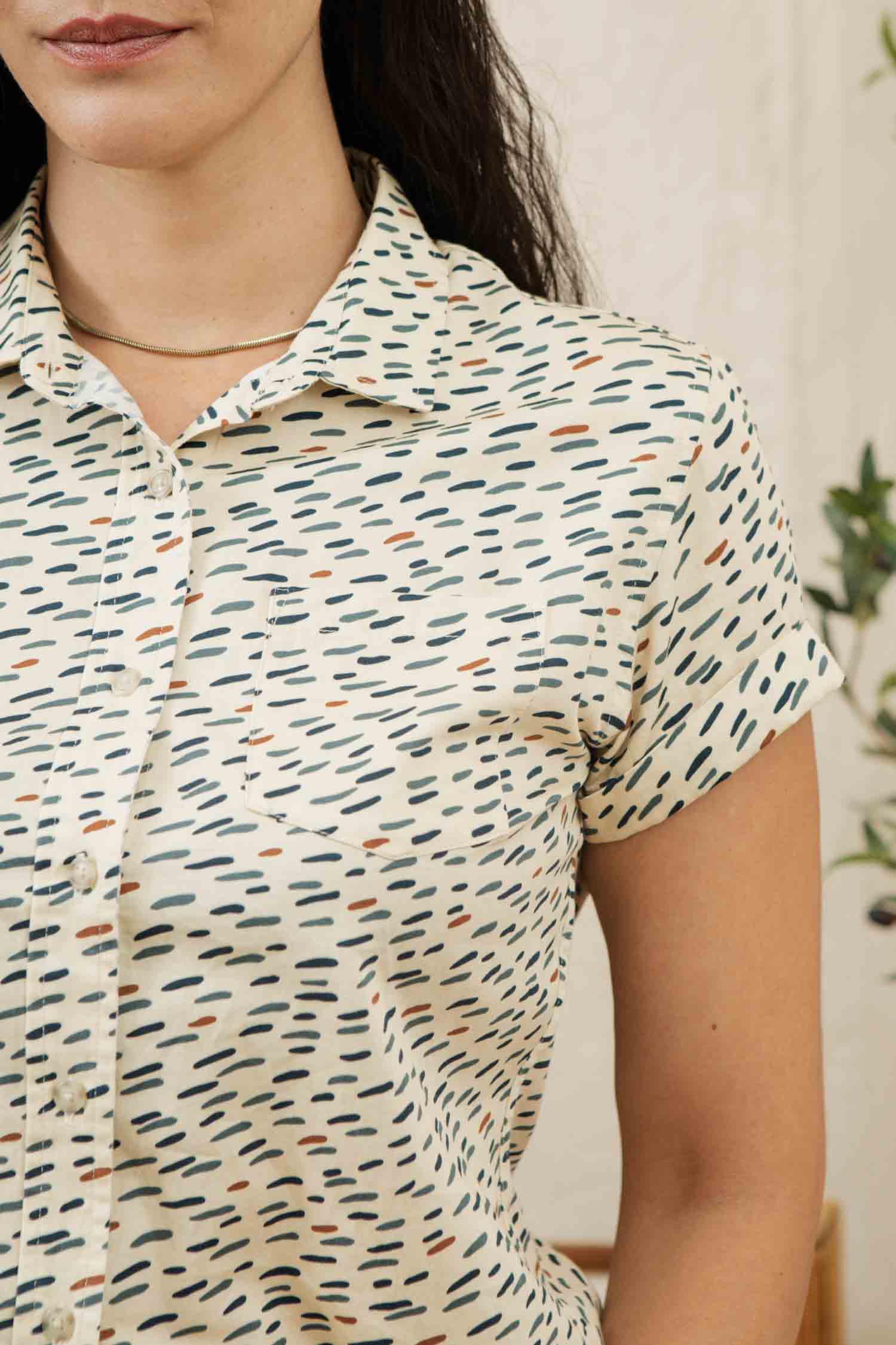 A woman wears the Bridge & Burn Bea Shirt in Ivory Dashes, a classic short-sleeve button-down with black and brown dashes on light cream organic cotton. Only her upper body and part of her face are visible.