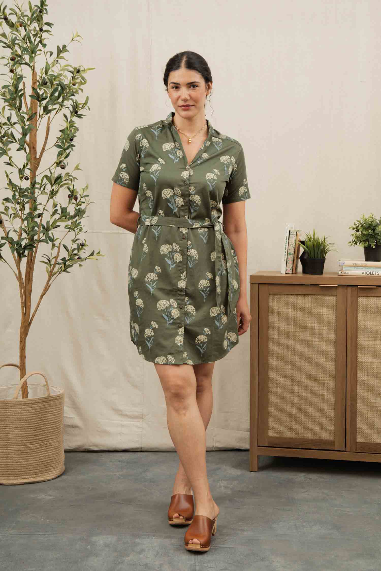 The Sofia Shirt Dress by Bridge & Burn is a short-sleeve, button-front dress in olive green organic cotton, featuring a belted waist and a white and blue wildflower print with green leaves. The dress is shown flat against a plain background.