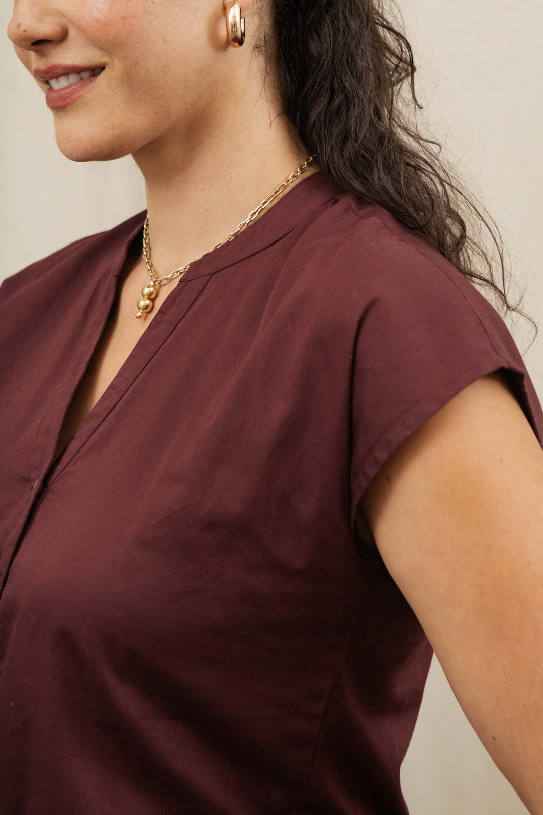 A woman with long dark hair wears the Bridge & Burn Ladd Shirt in burgundy, along with a gold chain pendant necklace and gold hoop earrings. She is smiling, with only her upper torso and side of her face visible.