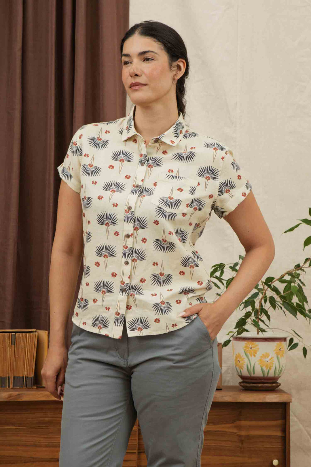 A woman with dark hair tied back wears the Bridge & Burn Bea Shirt in Aloha Floral, a short-sleeve button-down linen cotton shirt, and gray pants. She stands indoors by a plant and wooden furniture with one hand in her pocket.