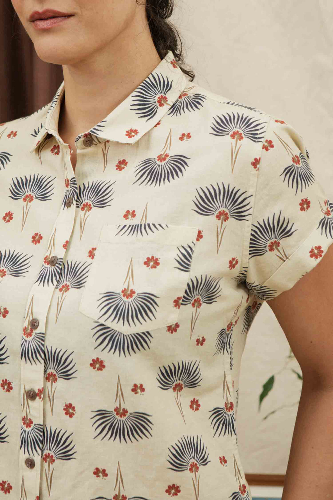 A woman models the Bridge & Burn Bea Shirt / Aloha Floral, a cream short-sleeve button-down with collar and pocket, featuring blue fan-shaped leaves and small red flowers. Only her lower face and upper torso are shown.