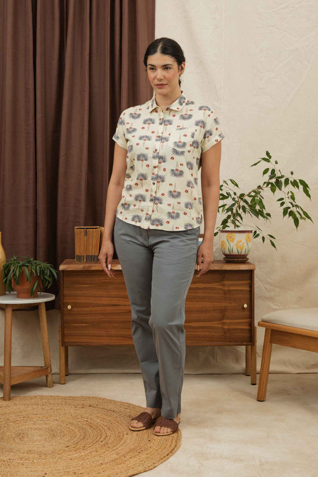 A woman stands indoors on a woven rug, wearing the Bridge & Burn Bea Shirt in Aloha Floral, paired with gray pants and brown sandals. Behind her are a wooden sideboard, chairs, potted plants, and brown curtains.