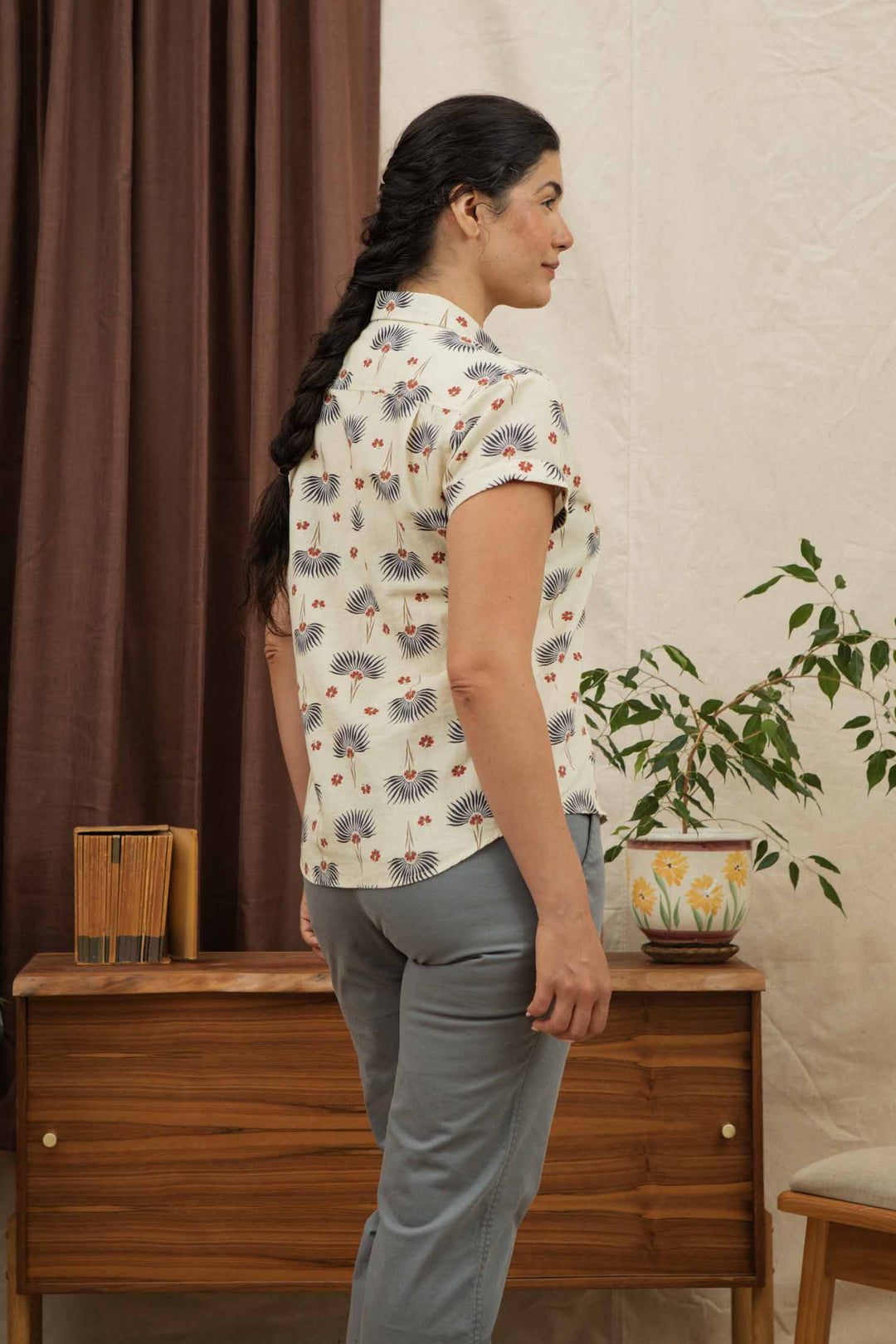 A woman with long dark hair in a braid stands sideways wearing the Bridge & Burn Bea Shirt in Aloha Floral, paired with gray pants. Behind her are a wooden cabinet, books, a potted plant, and brown curtains.