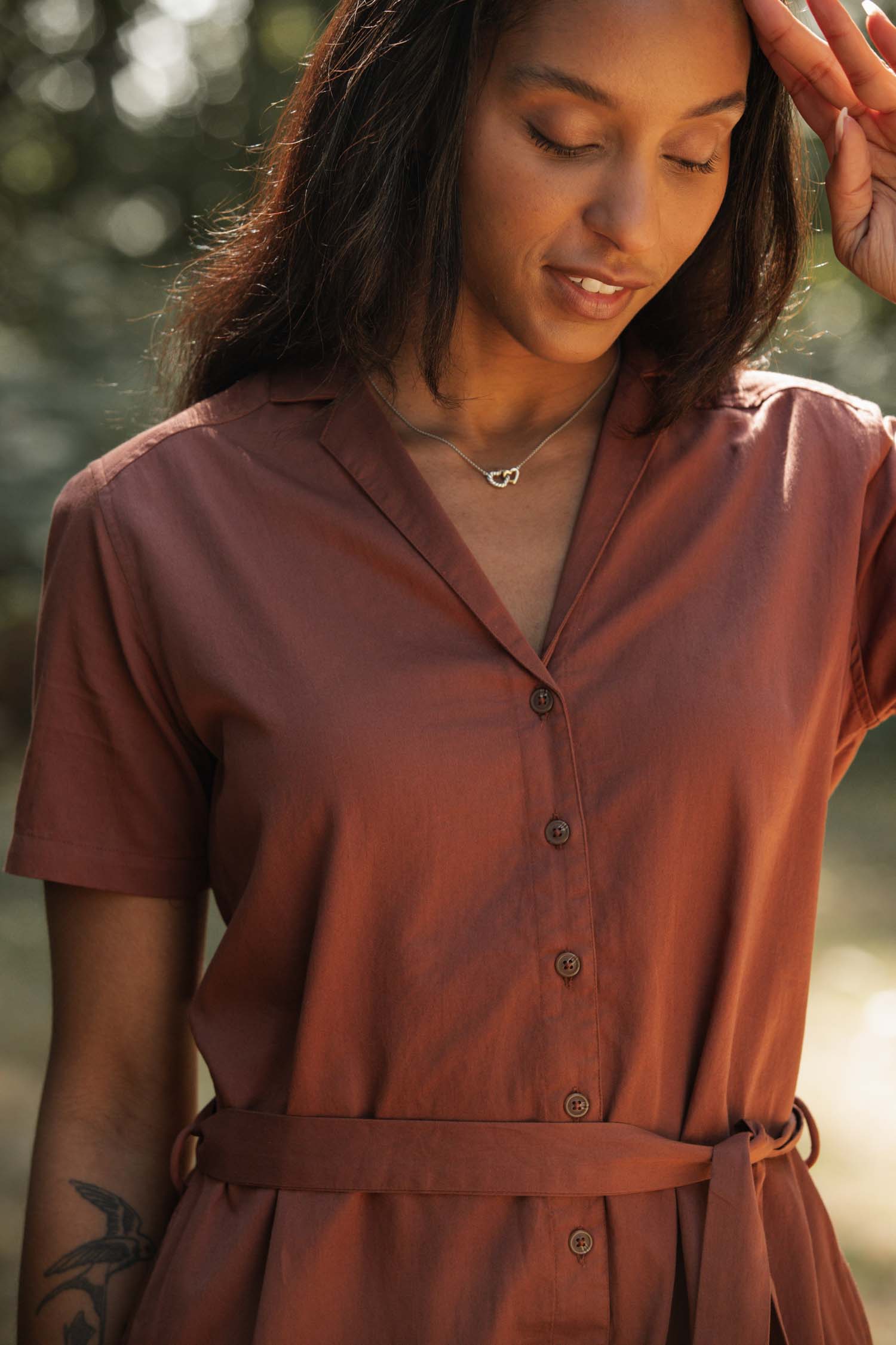 A woman in the Bridge & Burn Sofia Shirt Dress in Copper stands outdoors, smiling gently with one hand to her forehead as sunlight filters through the trees, highlighting the dress’s lightweight design.