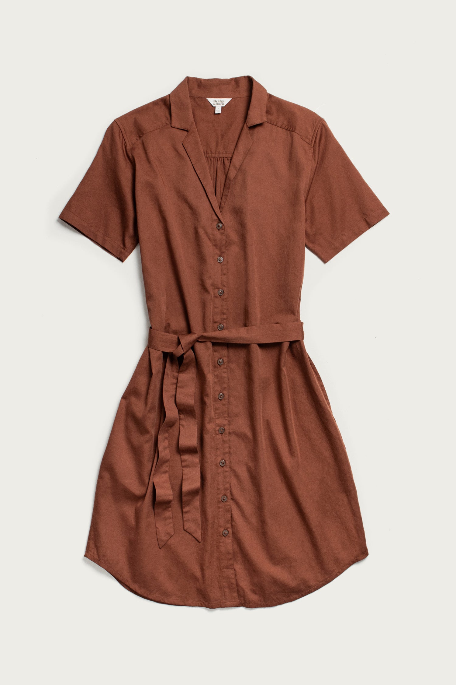 The Bridge & Burn Sofia Shirt Dress in Copper is a lightweight, short-sleeved button-front dress with a collar and matching fabric belt, crafted from rust-brown organic cotton and shown laid flat on a light background.