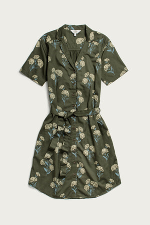The Sofia Shirt Dress by Bridge & Burn is a short-sleeve, button-front dress in olive green organic cotton, featuring a belted waist and a white and blue wildflower print with green leaves. The dress is shown flat against a plain background.