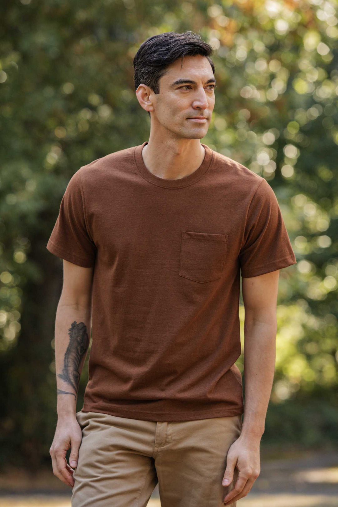 A man with short dark hair and a tattoo on his left forearm stands outdoors among green trees, wearing the Bridge & Burn Recycled Pocket Tee in Bronze with a chest pocket, paired with beige pants. He looks slightly to the side.