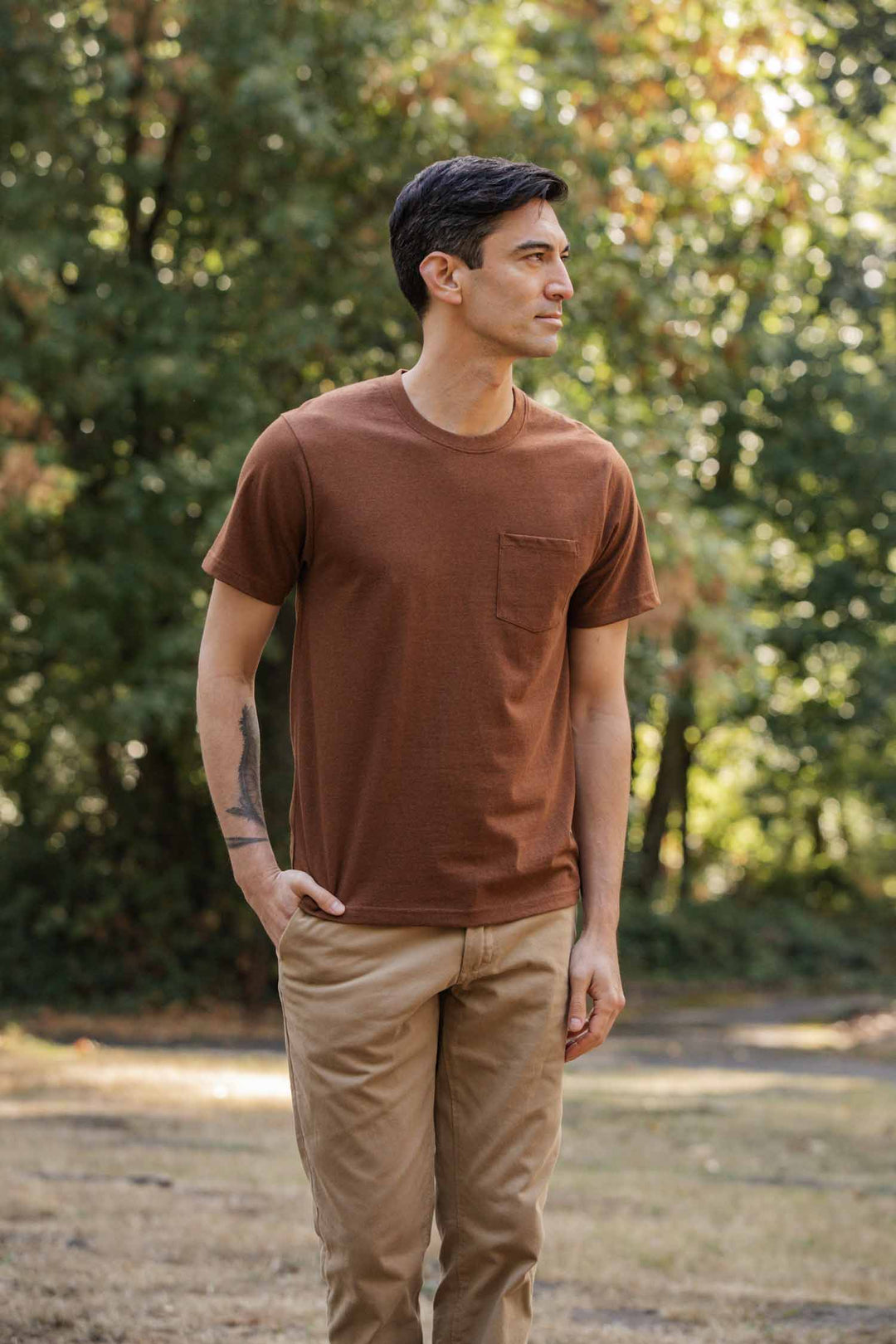 A man stands in a sunlit park wearing the Bridge & Burn Recycled Pocket Tee in Bronze and tan pants. He has short dark hair, a tattoo on his left forearm, and looks to his left with one hand in his pocket. Trees fill the background.