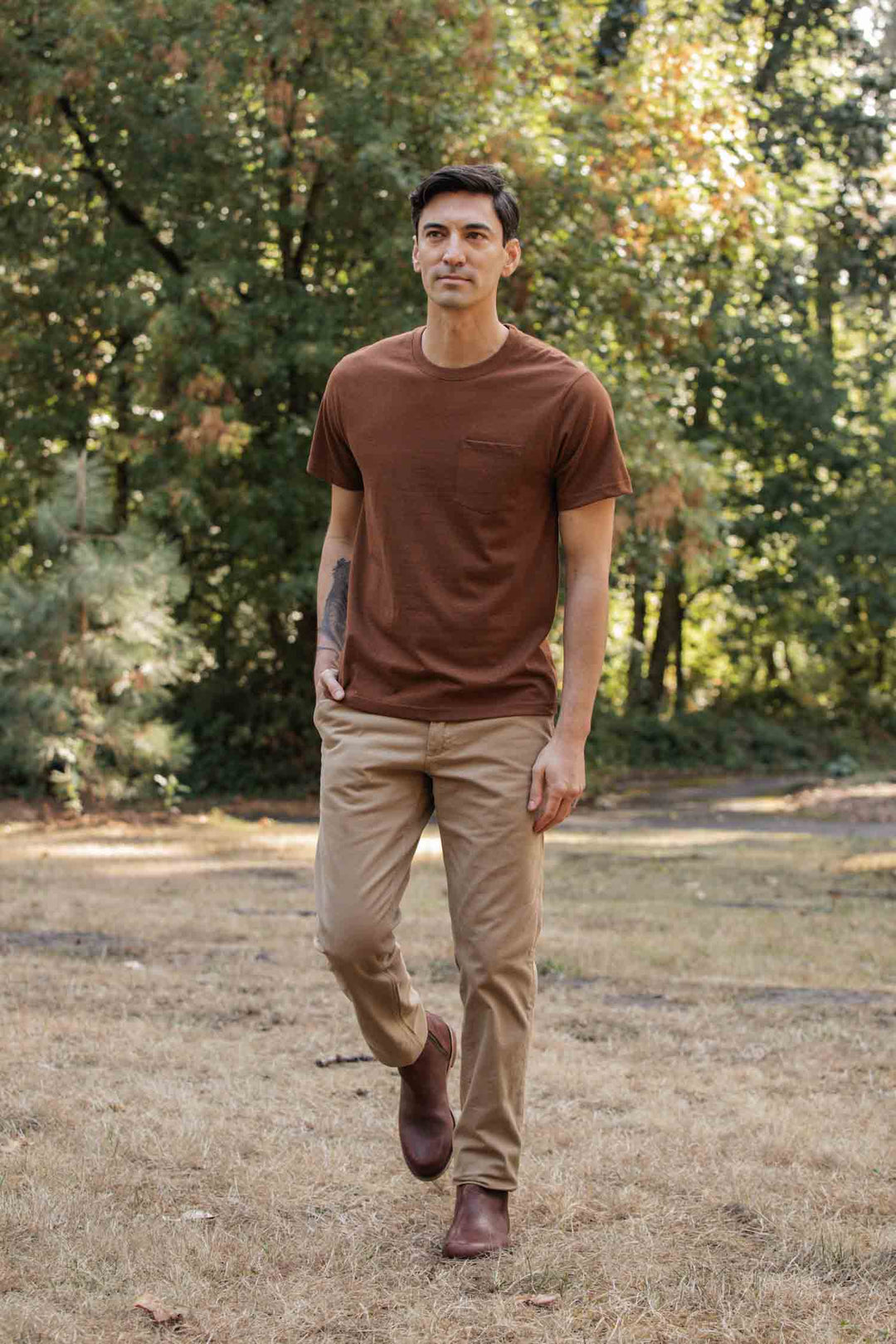 A man wearing the Bridge & Burn Recycled Pocket Tee in Bronze, tan pants, and brown shoes walks outdoors on dry grass with green trees in the background.