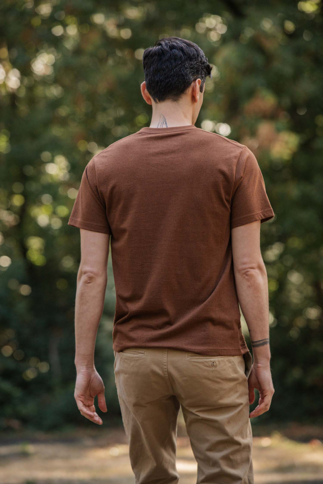 A person with short dark hair stands outdoors facing away, wearing the Bridge & Burn Recycled Pocket Tee in Bronze. A neck tattoo is visible, and blurred trees and greenery make up the background.