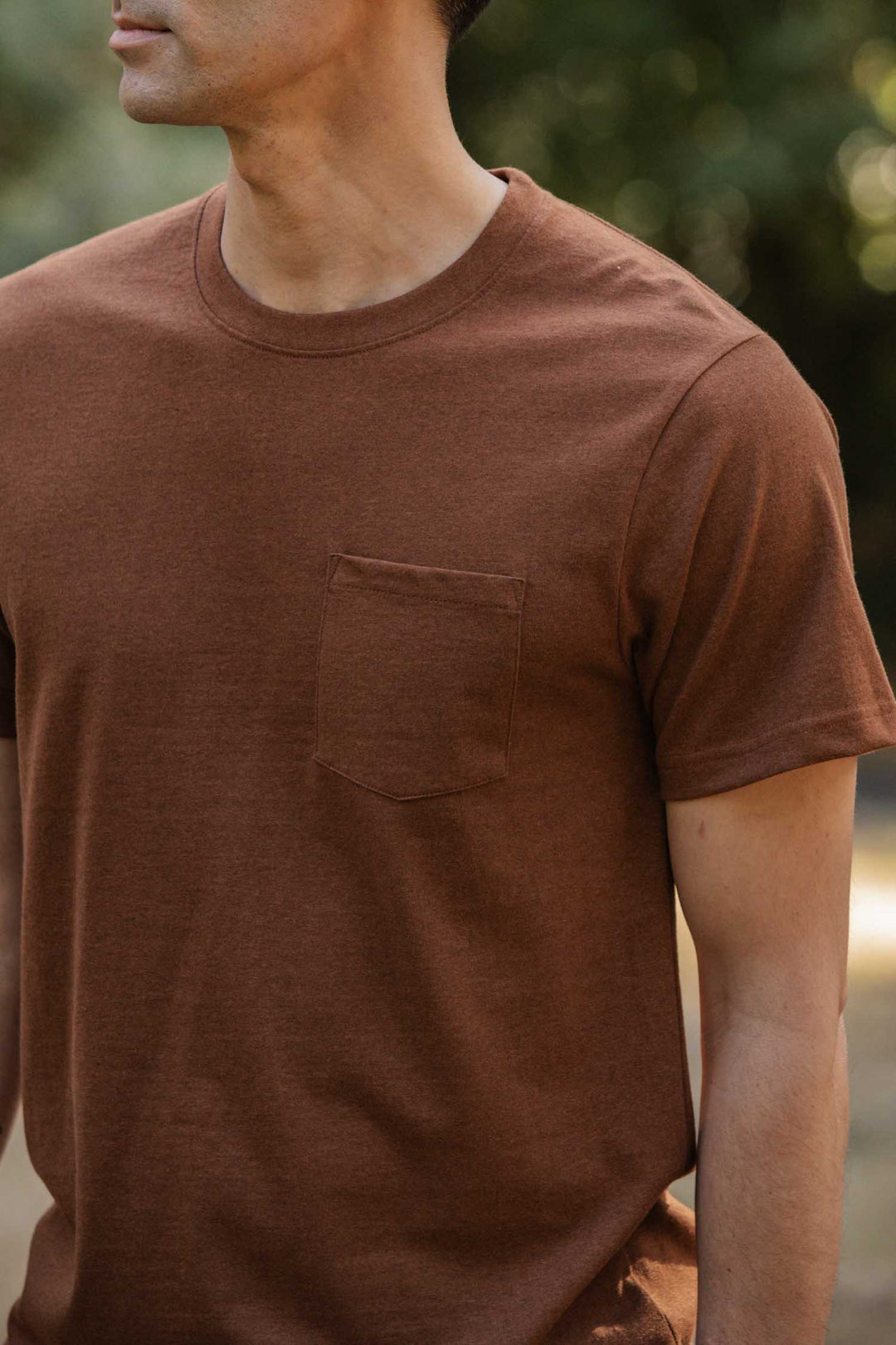 A man stands outdoors in the Bridge & Burn Recycled Pocket Tee in Bronze. The short-sleeve t-shirt features a chest pocket. Greenery forms a blurred background; his face is partially out of frame.