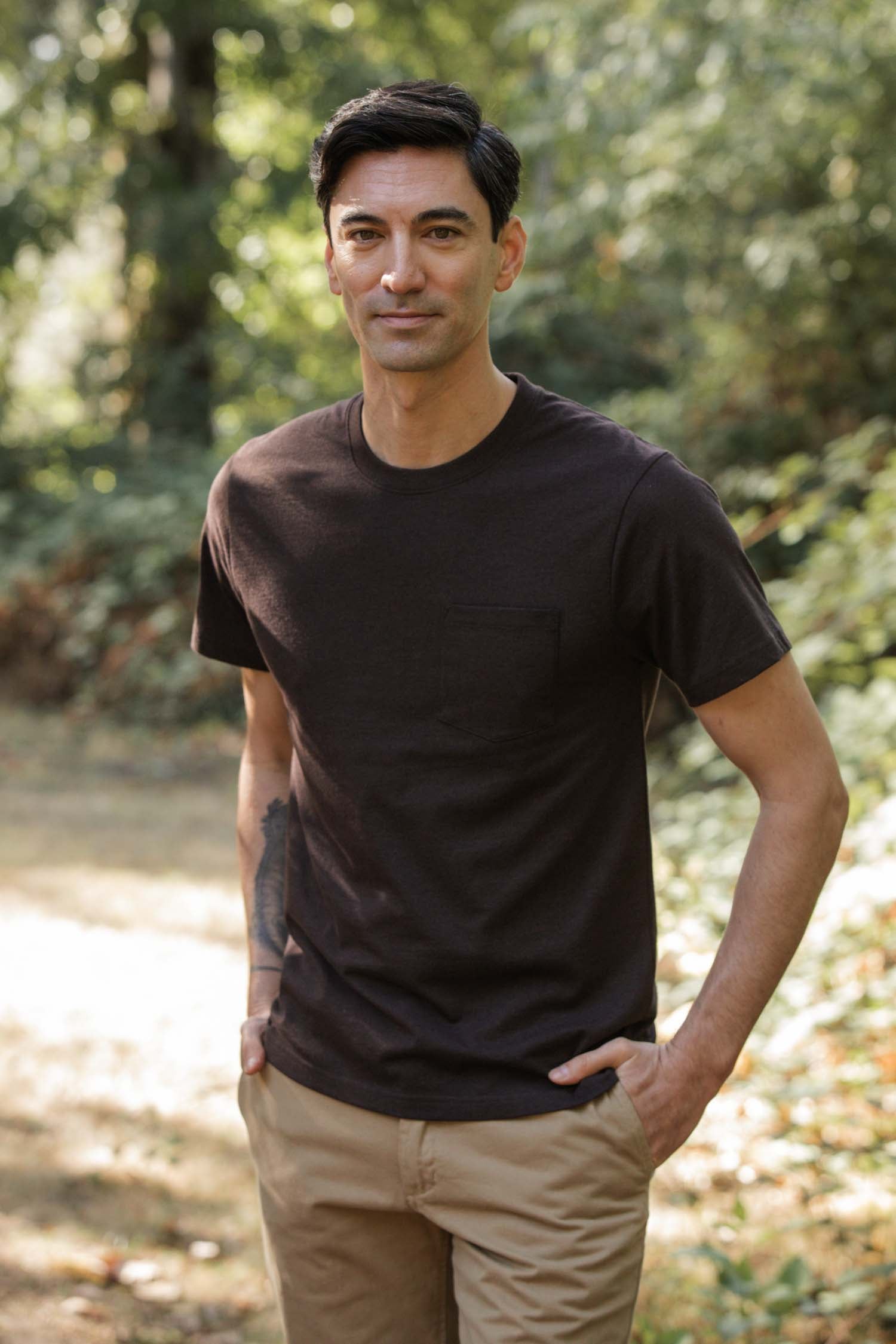 A man with short dark hair stands outdoors in the sun, smiling slightly at the camera. He wears the Bridge & Burn Recycled Pocket Tee in Truffle and tan pants, hands in his pockets.