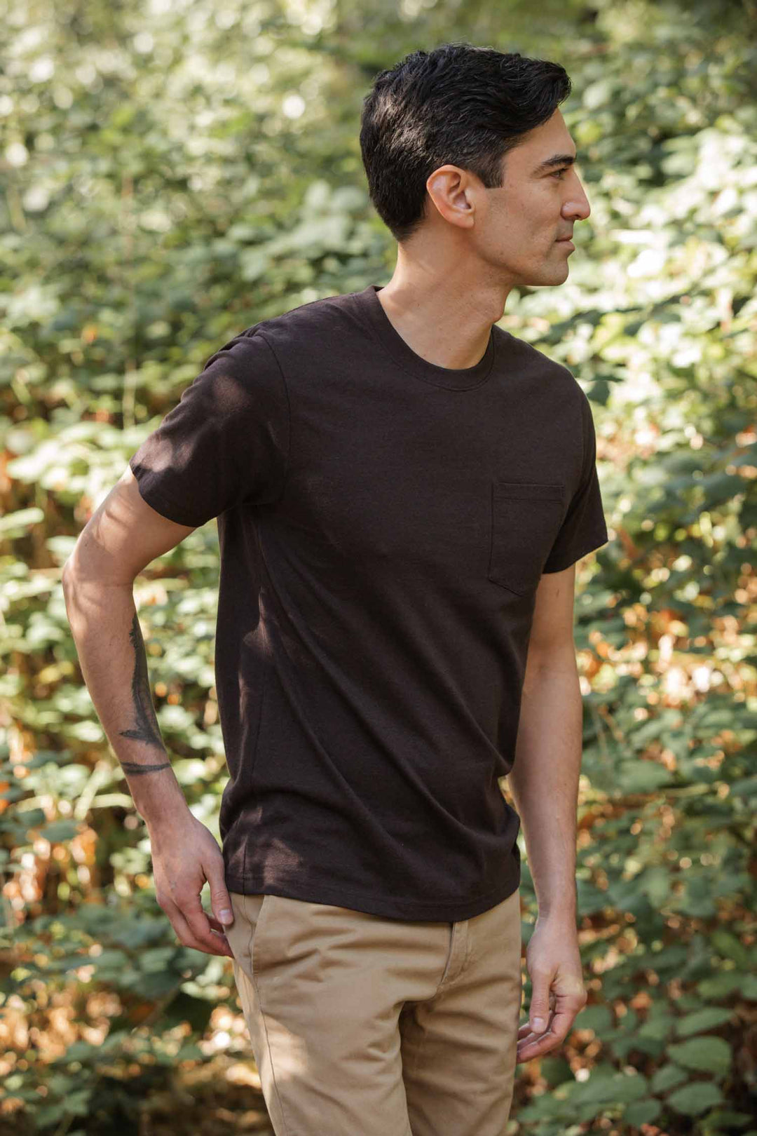 A man with short dark hair stands outdoors by green plants, wearing the Bridge & Burn Recycled Pocket Tee in Truffle and beige pants. He looks to his right with a neutral expression.