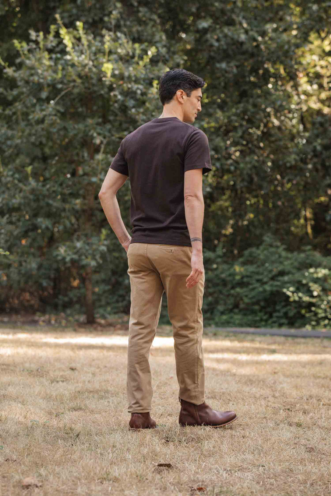 A man stands outdoors on dry grass, facing away from the camera. He wears Bridge & Burn’s Recycled Pocket Tee in Truffle, with khaki pants and brown boots. Green trees fill the background.
