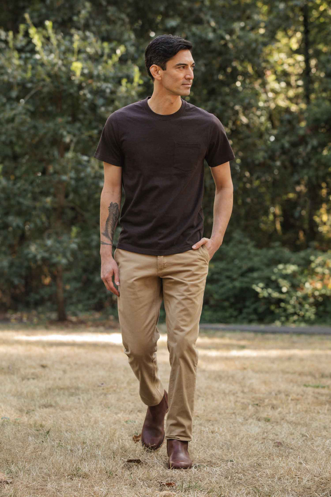 A man with short dark hair wears the Bridge & Burn Recycled Pocket Tee in Truffle, tan pants, and brown shoes as he walks on dry grass in a park with green trees. One hand is in his pocket and he looks to the side.