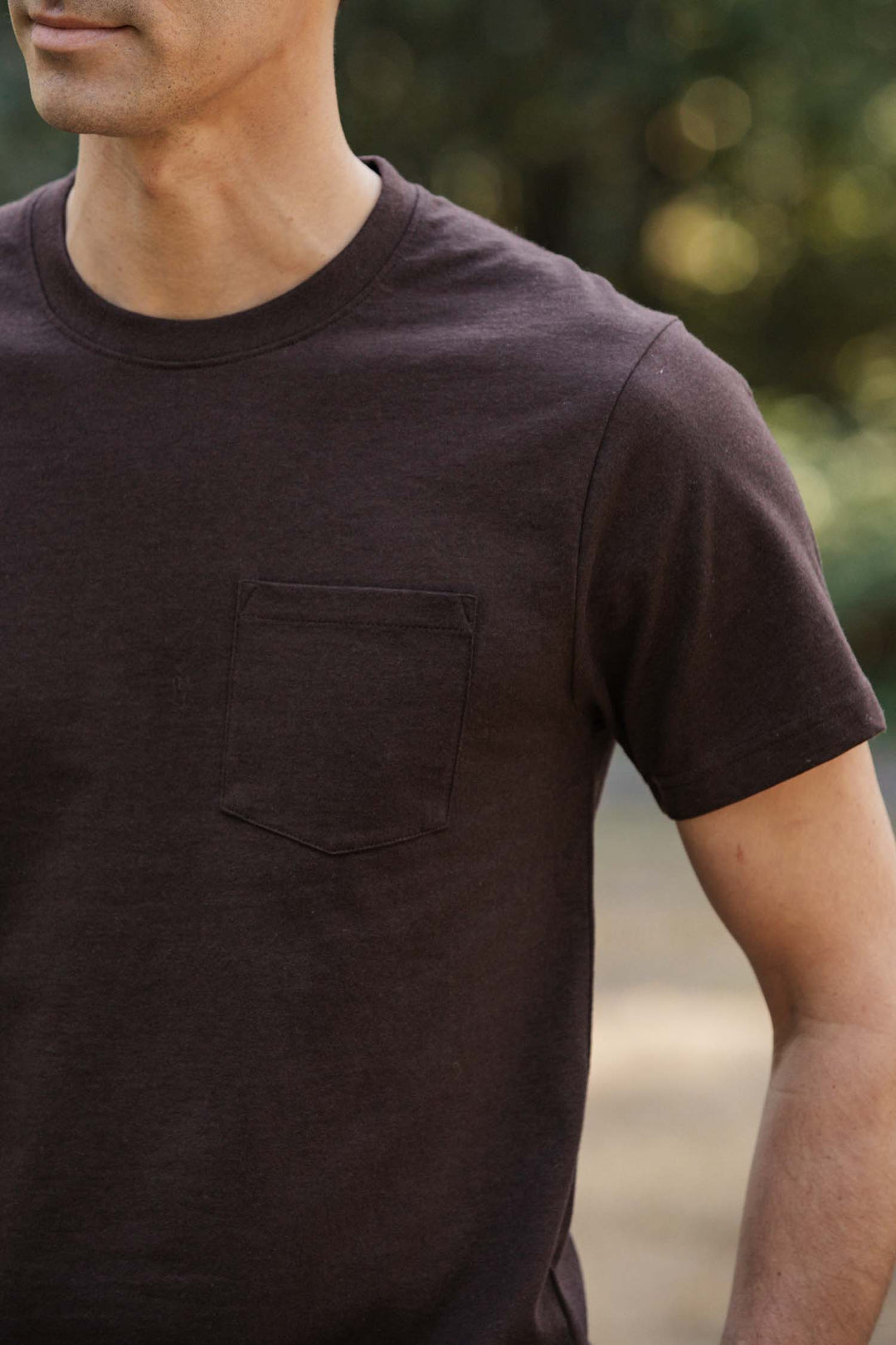 A man wears the Bridge & Burn Recycled Pocket Tee in Truffle, a dark brown short-sleeve shirt with a chest pocket, standing outdoors with blurred greenery in the background. Only his upper body is visible.