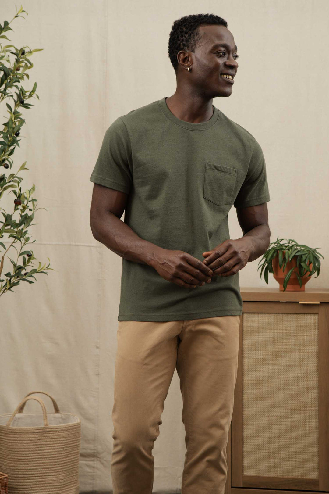 A man stands indoors, smiling, wearing the Bridge & Burn Recycled Pocket Tee in Olive and beige pants. Potted plants and wicker baskets surround him against a neutral wall.