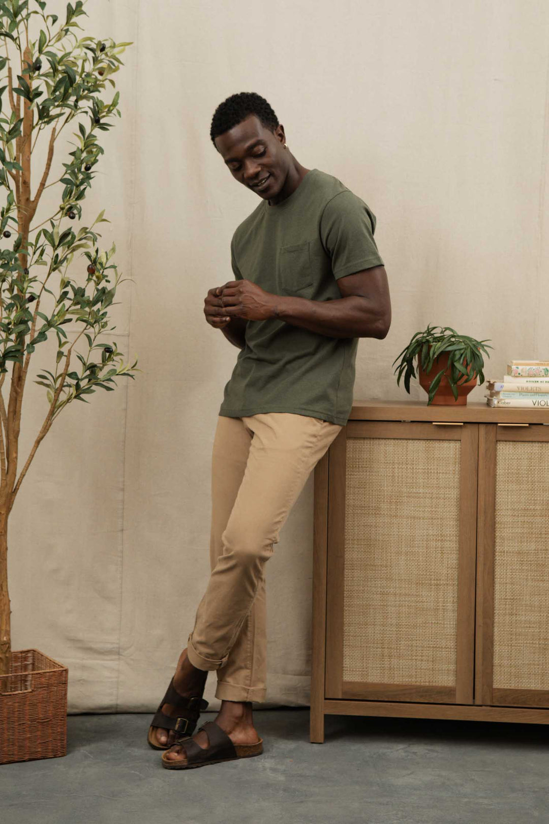 A man in the Bridge & Burn Recycled Pocket Tee in Olive and beige pants leans casually against a cabinet with books and a potted plant, smiling next to a tall indoor plant in a basket.