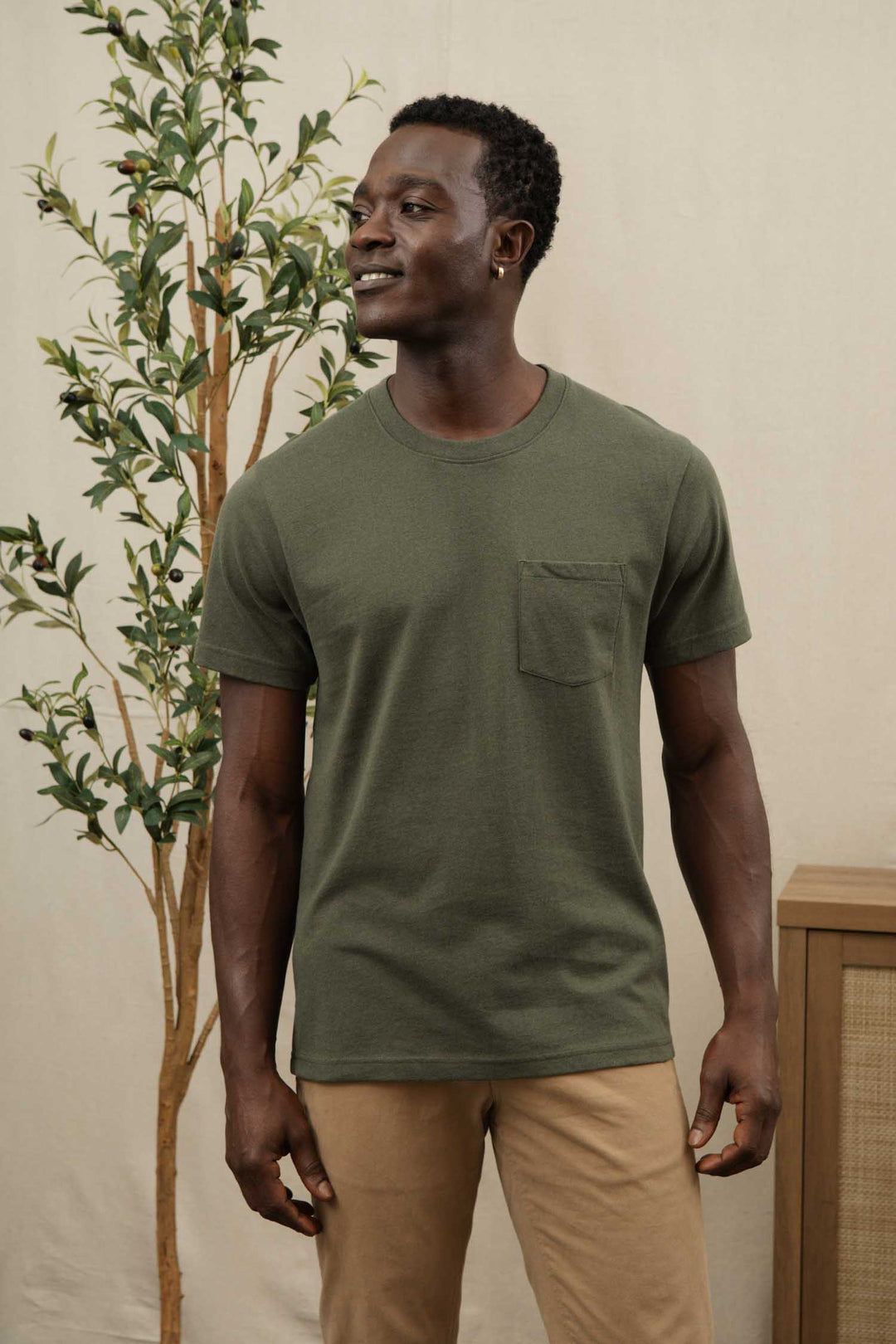 A man wearing the Bridge & Burn Recycled Pocket Tee in olive and beige pants stands indoors near a leafy plant, smiling and looking slightly left. The neutral background features a partially visible wooden furniture piece.