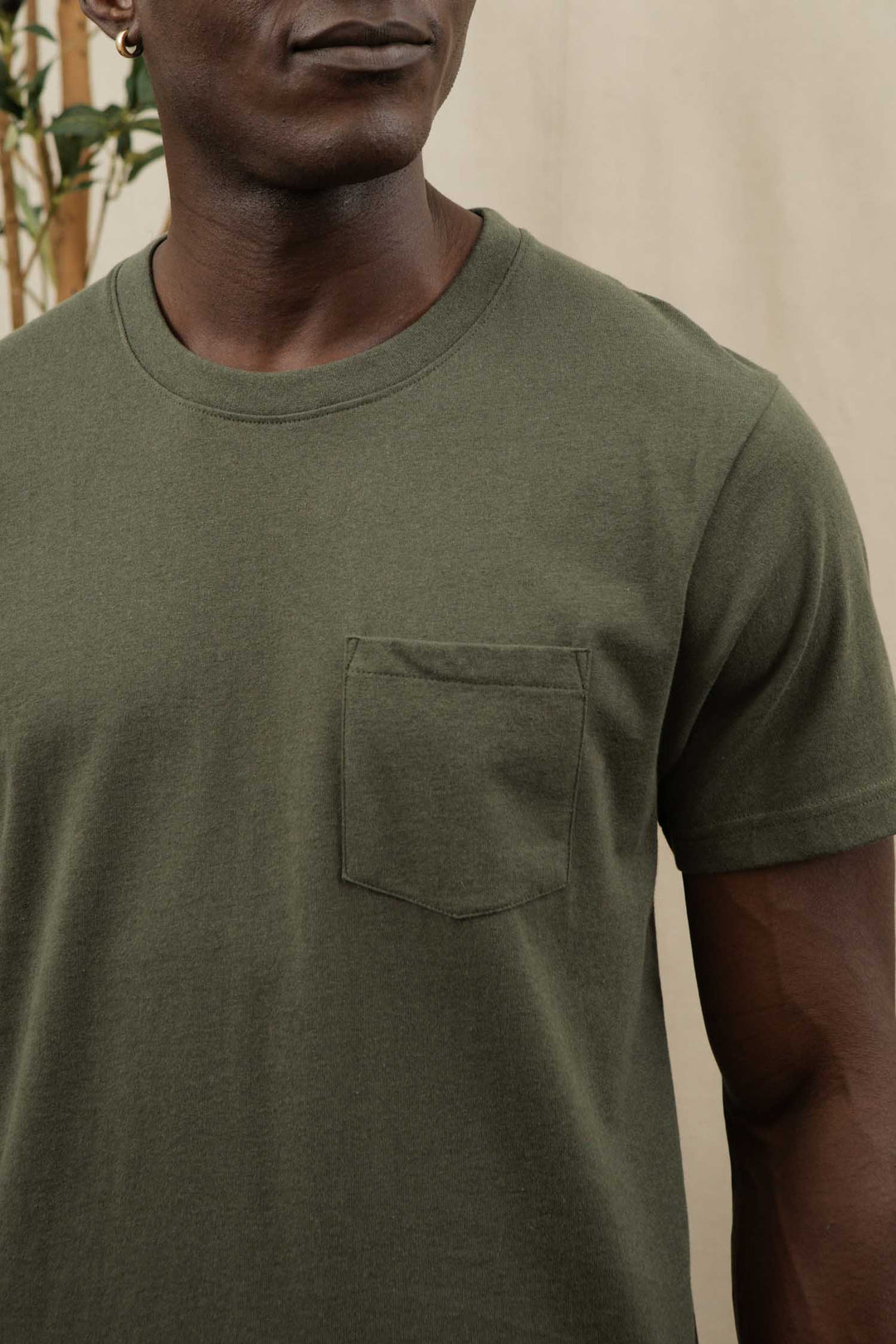 A person wearing the Bridge & Burn Recycled Pocket Tee in olive stands indoors. The cropped image highlights the sustainable crew neck with chest pocket, showing just the upper torso and part of the face.