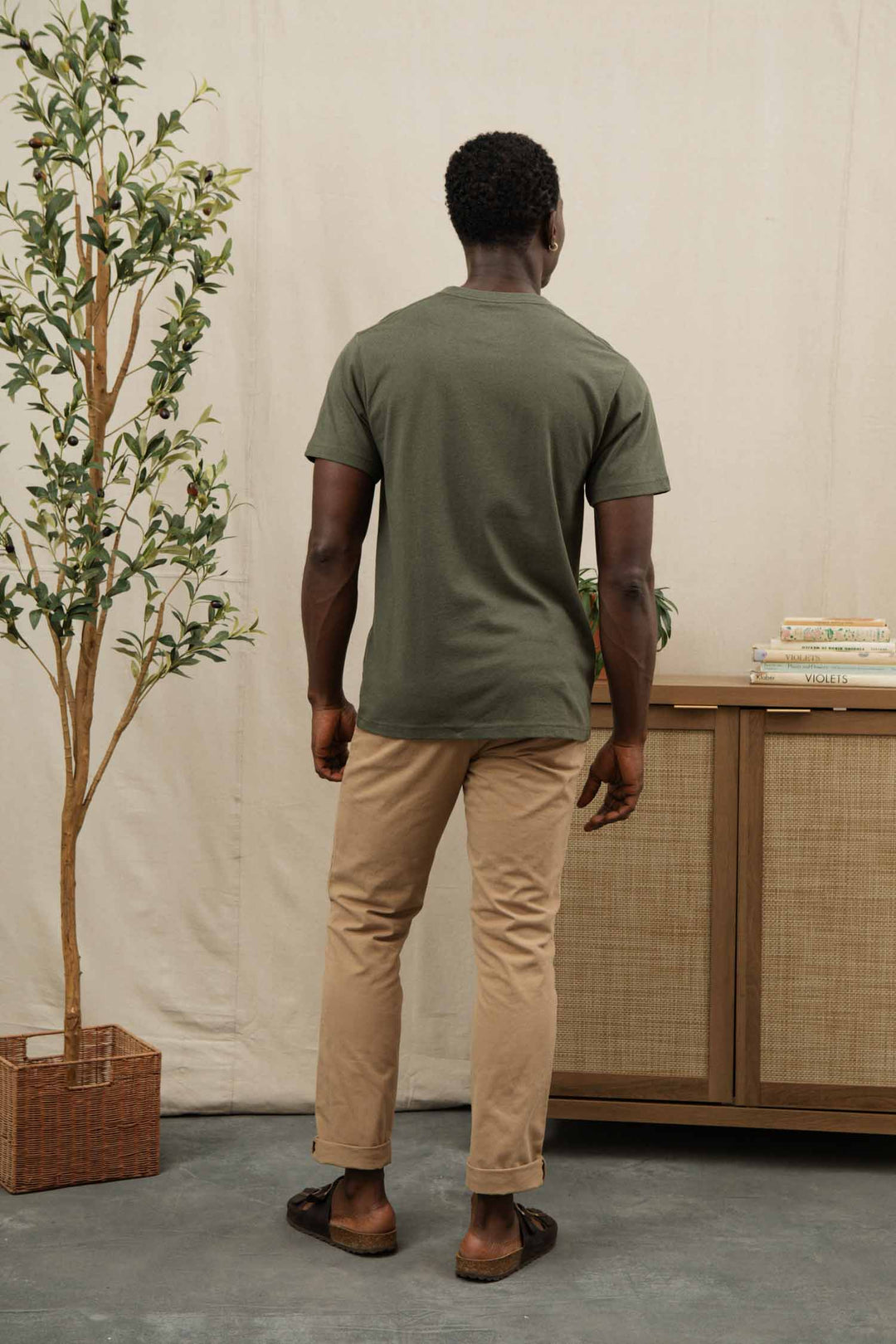 A man stands indoors with his back to the camera, wearing the Bridge & Burn Recycled Pocket Tee in Olive, beige pants, and brown shoes. Beside him are a potted plant and a wooden cabinet stacked with books.