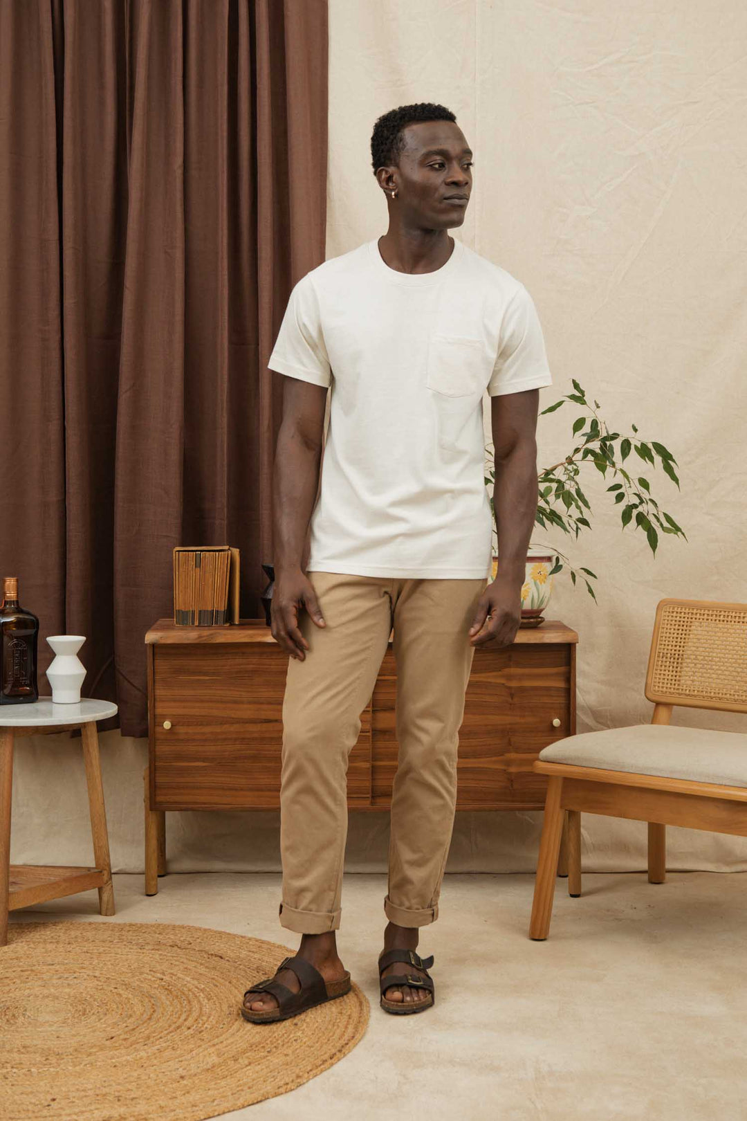 A man stands indoors wearing the Bridge & Burn Recycled Pocket Tee in Ivory, paired with beige pants and dark sandals. He looks to his left, surrounded by furniture, a potted plant, and neutral-toned decor.