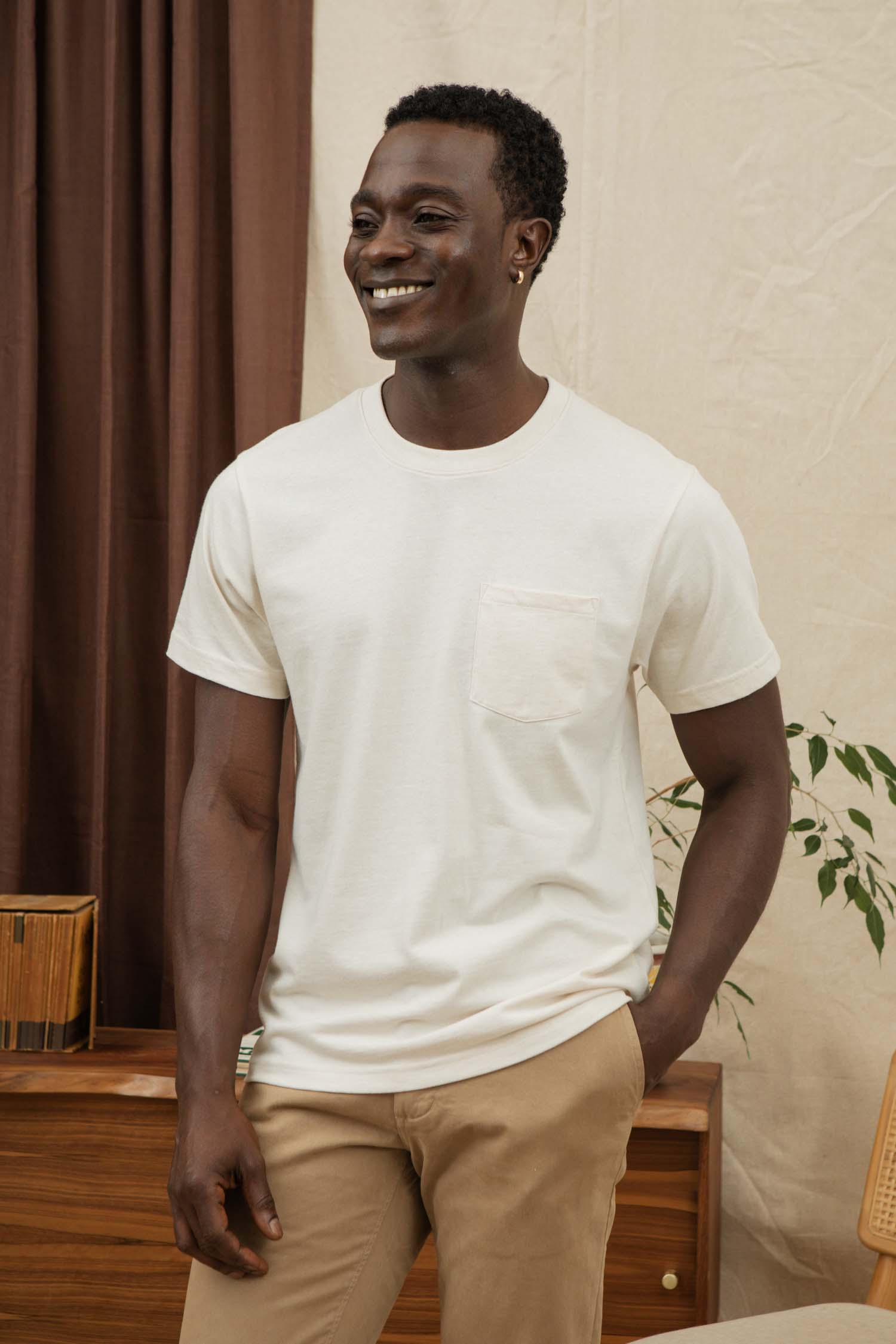 A man wears the Bridge & Burn Recycled Pocket Tee in Ivory with beige pants, standing indoors and smiling with one hand in his pocket. Behind him are a wooden sideboard, a plant, and brown and cream curtains.
