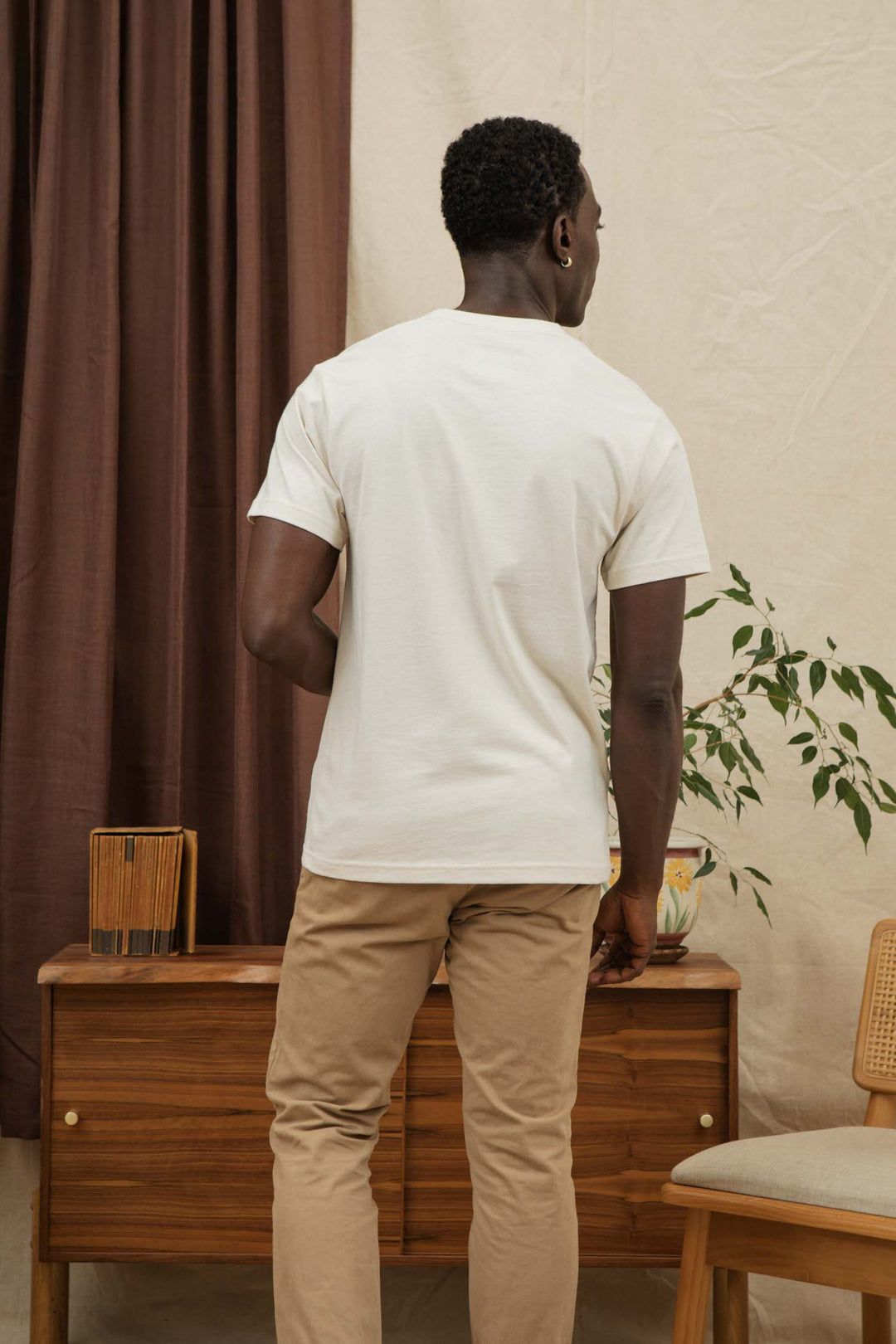 A man wearing the Bridge & Burn Recycled Pocket Tee in Ivory and beige pants stands with his back to the camera in a room featuring a wooden dresser, chair, potted plant, and brown curtains.