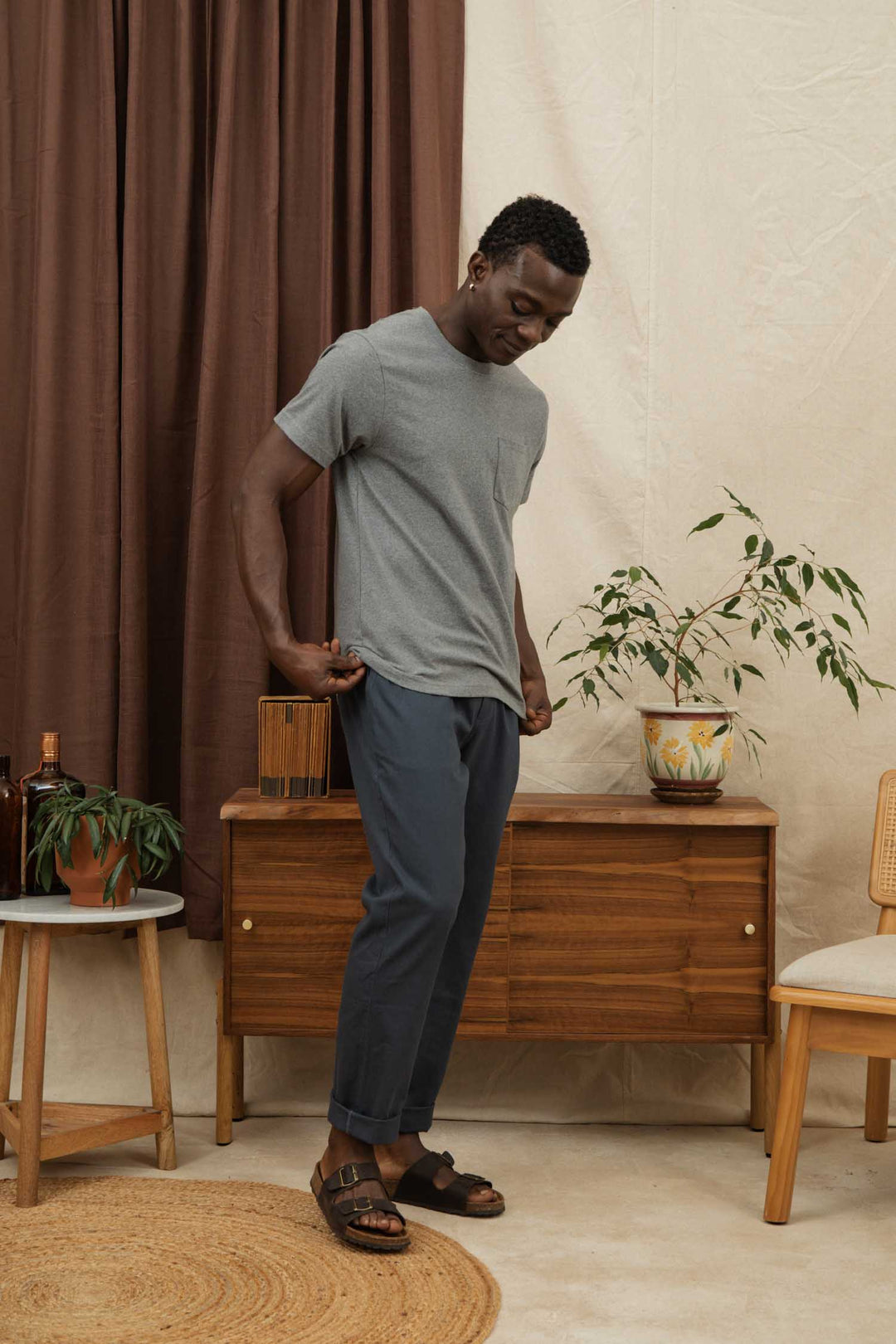 A man stands indoors on a round rug, wearing the Bridge & Burn Recycled Pocket Tee in Grey Heather, navy pants, and brown sandals. Behind him are a wooden cabinet, potted plants, a stool, a chair, and brown curtains.