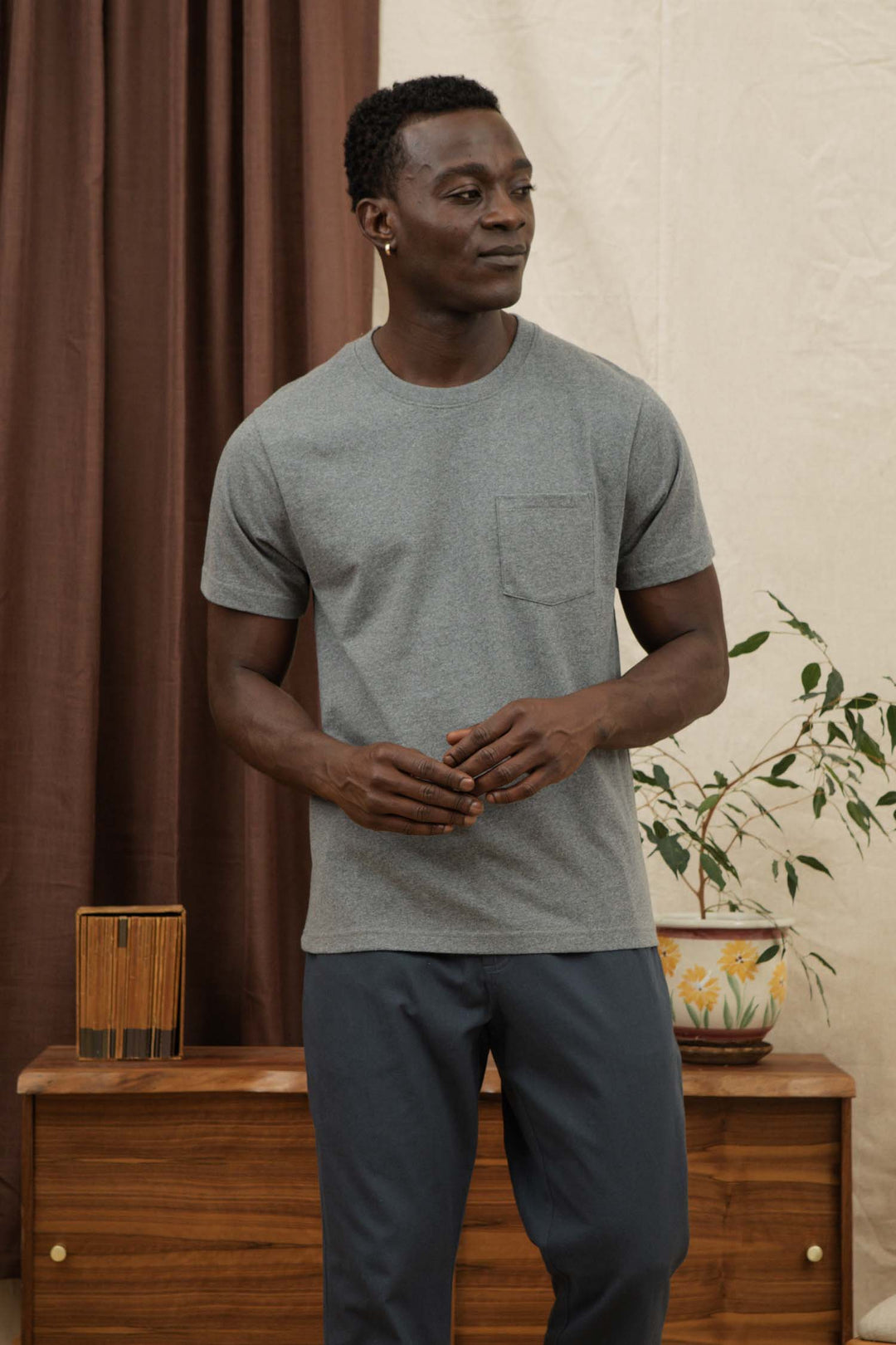 A man wears the Bridge & Burn Recycled Pocket Tee in Grey Heather and dark pants indoors, standing by a wooden dresser with a potted plant and brown curtains in the background.