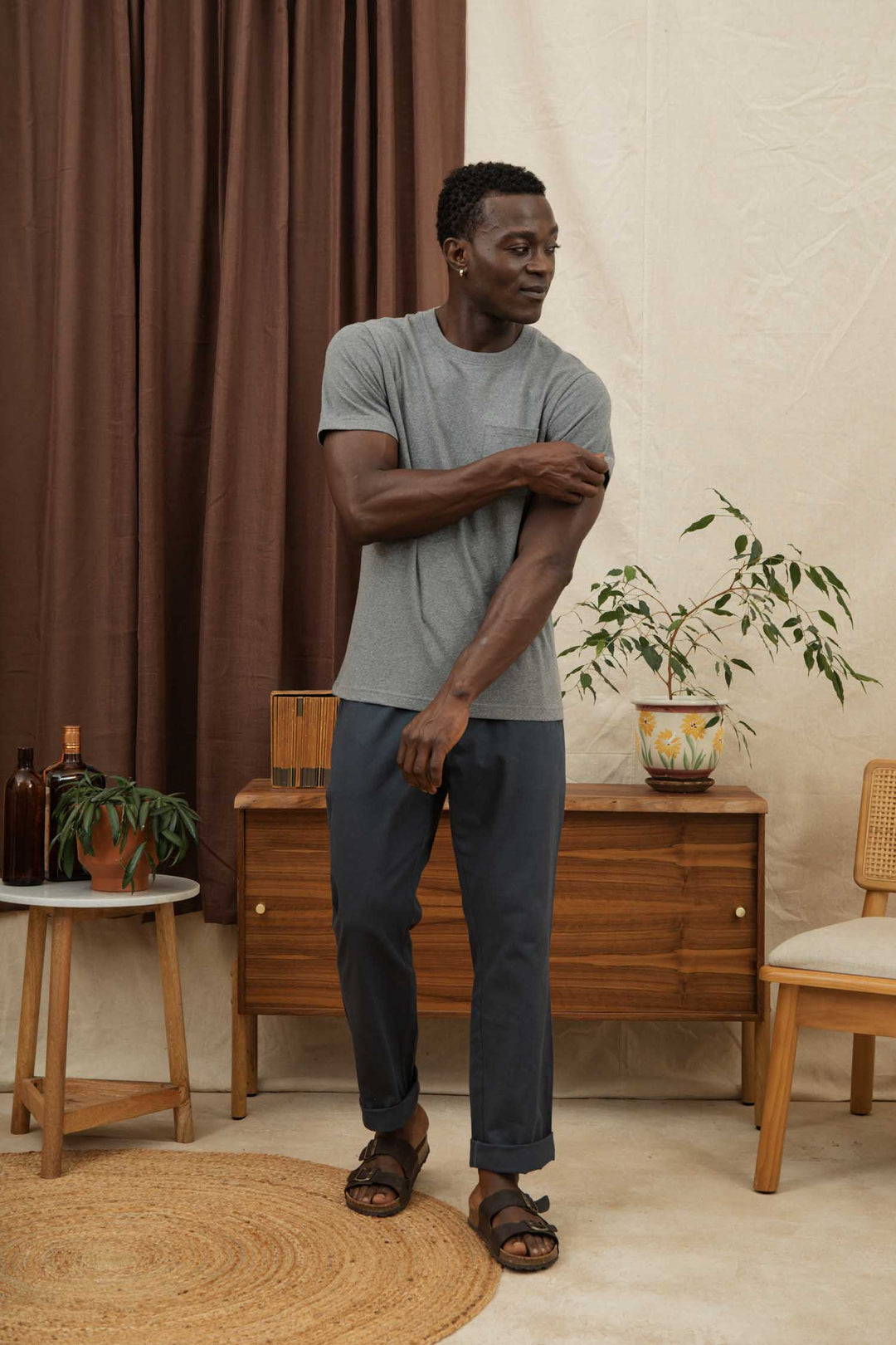A man adjusts his sleeve indoors, wearing the Bridge & Burn Recycled Pocket Tee in Grey Heather, dark pants, and sandals. The cozy, mid-century modern space features a wooden cabinet, potted plants, chairs, and brown curtains.
