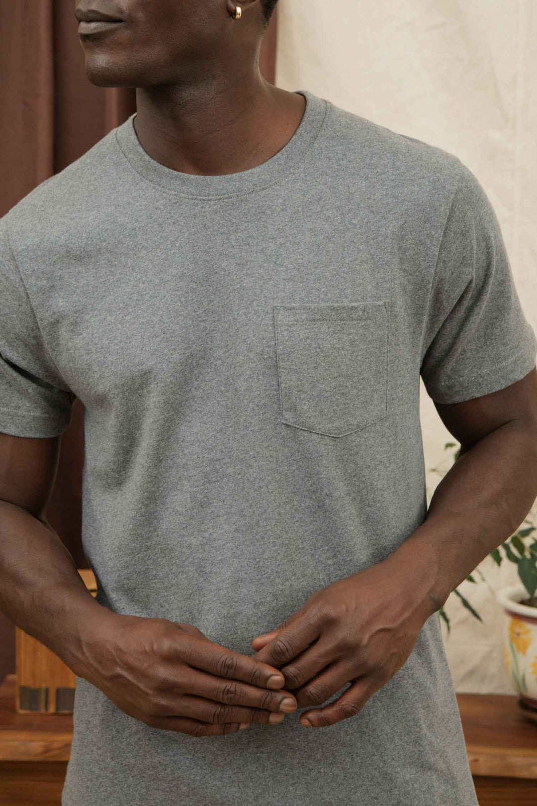 A man stands indoors, hands lightly clasped, wearing the Bridge & Burn Recycled Pocket Tee in Grey Heather. His face is partially out of frame.