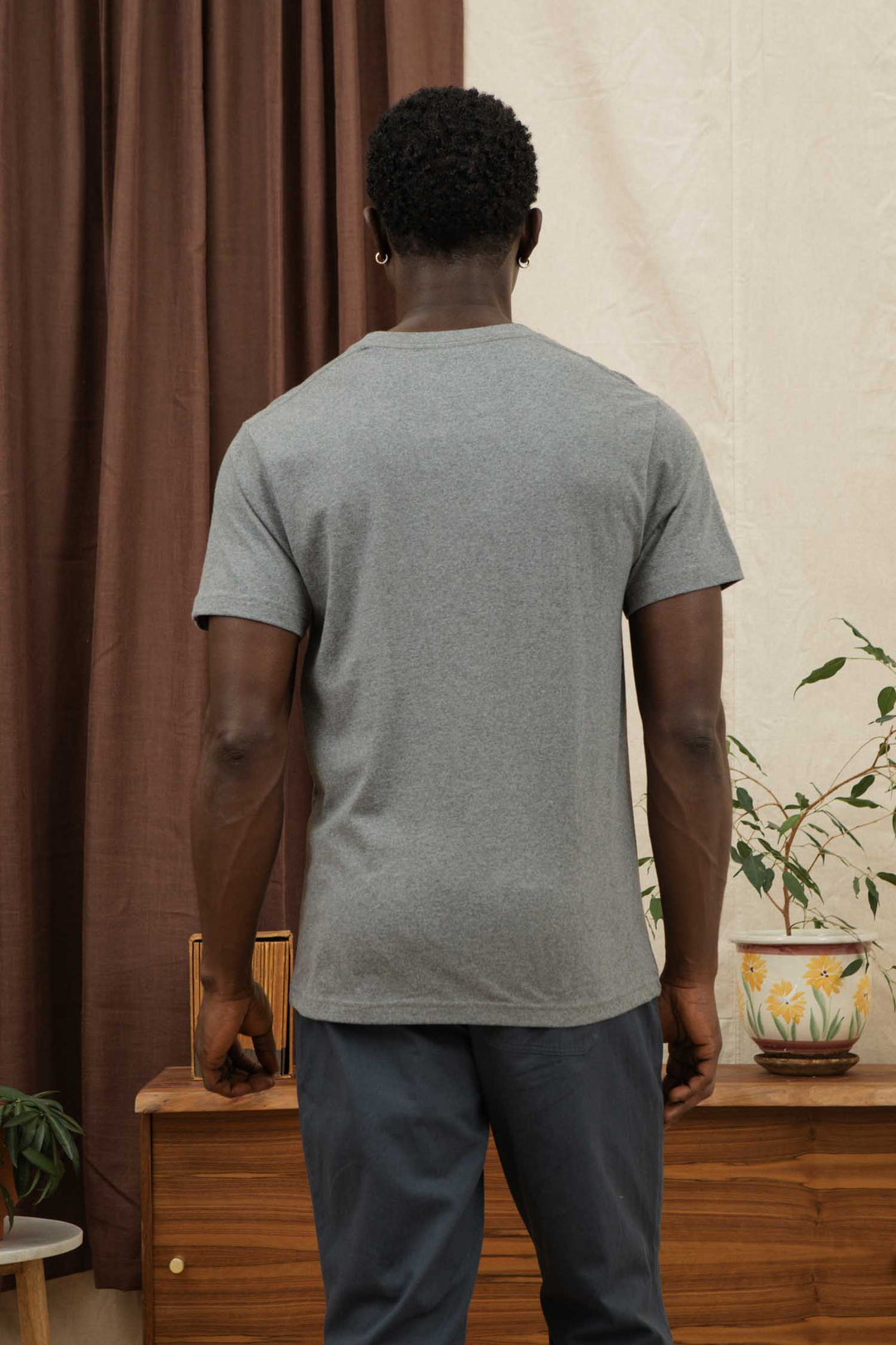 A man with short curly hair faces away, wearing the Bridge & Burn Recycled Pocket Tee in Grey Heather and dark pants, standing by a wooden cabinet with potted plants against brown curtains and a cream wall.