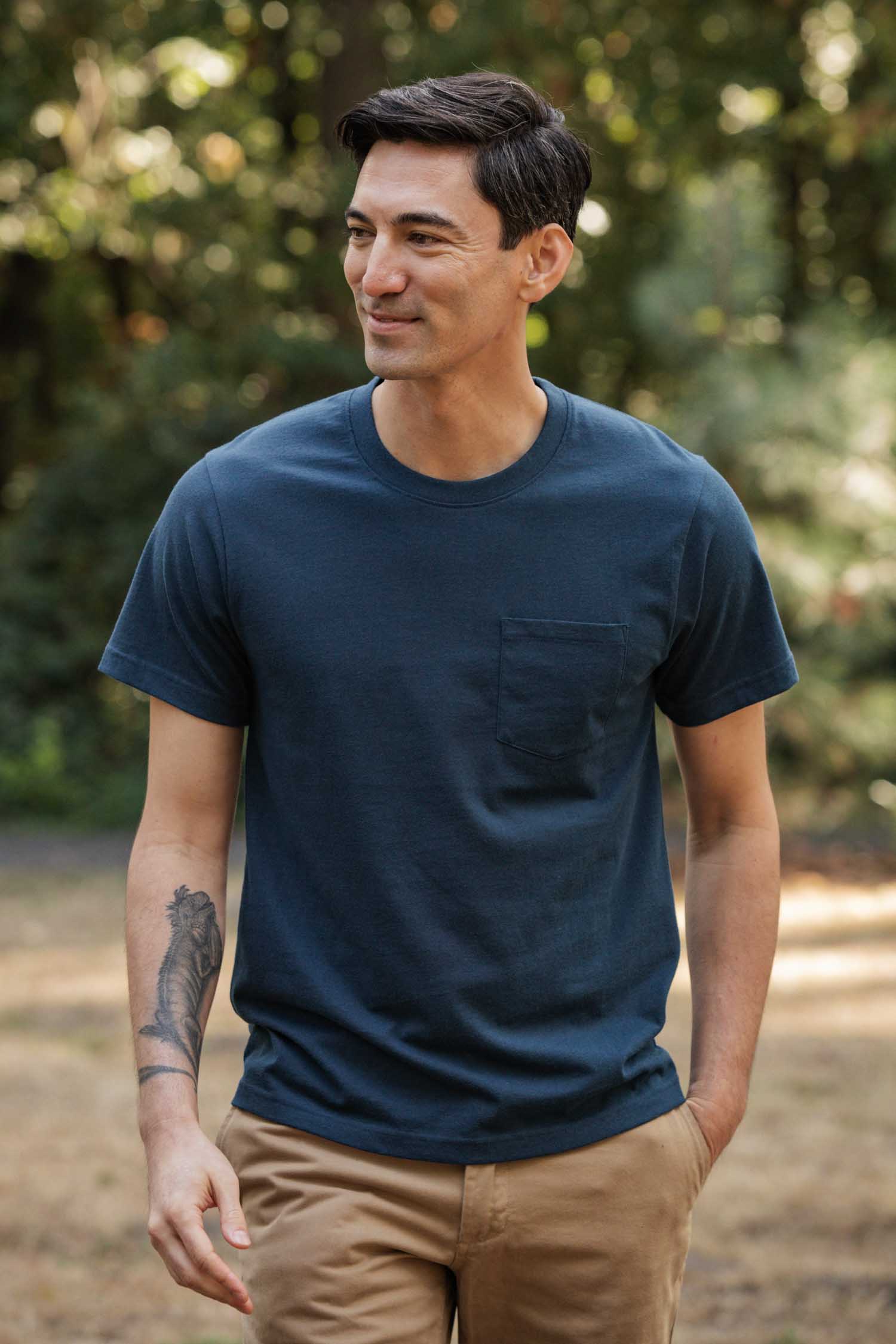 A man with short dark hair and a forearm tattoo stands outside among trees, smiling in tan pants and a Bridge & Burn Recycled Pocket Tee in Cobalt, featuring a chest pocket, for a vintage look.