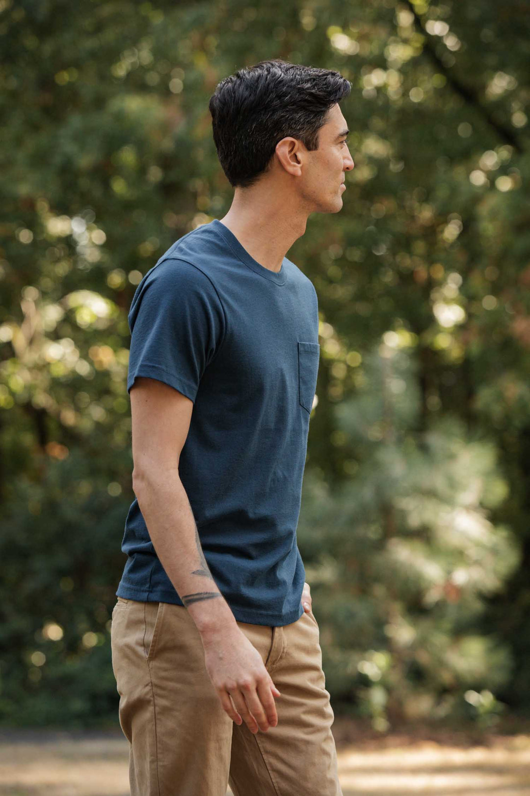A man in the Bridge & Burn Recycled Pocket Tee in Cobalt and tan pants stands outdoors, gazing right. With short dark hair and a tattooed left forearm, he’s set against leafy trees for a vintage vibe.