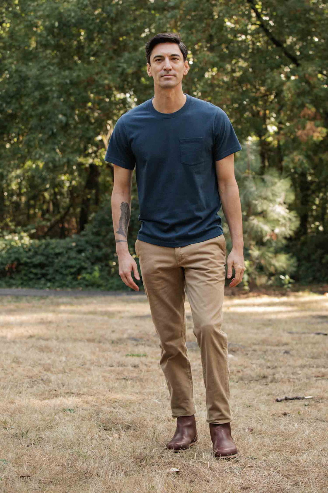 A man stands outdoors on dry grass, wearing the Bridge & Burn Recycled Pocket Tee in Cobalt with a vintage feel, paired with tan pants and brown boots. Trees and greenery are visible in the background.