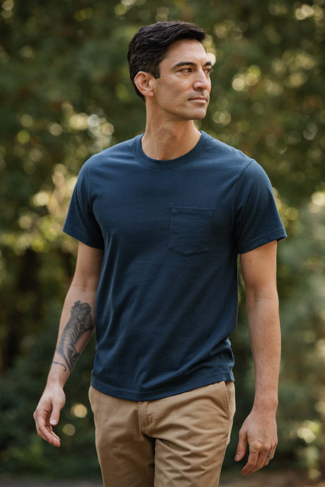 A man with short dark hair and a forearm tattoo stands outdoors among greenery, wearing tan pants and the Bridge & Burn Recycled Pocket Tee in Cobalt, giving the scene a subtle vintage feel as he looks to his left.