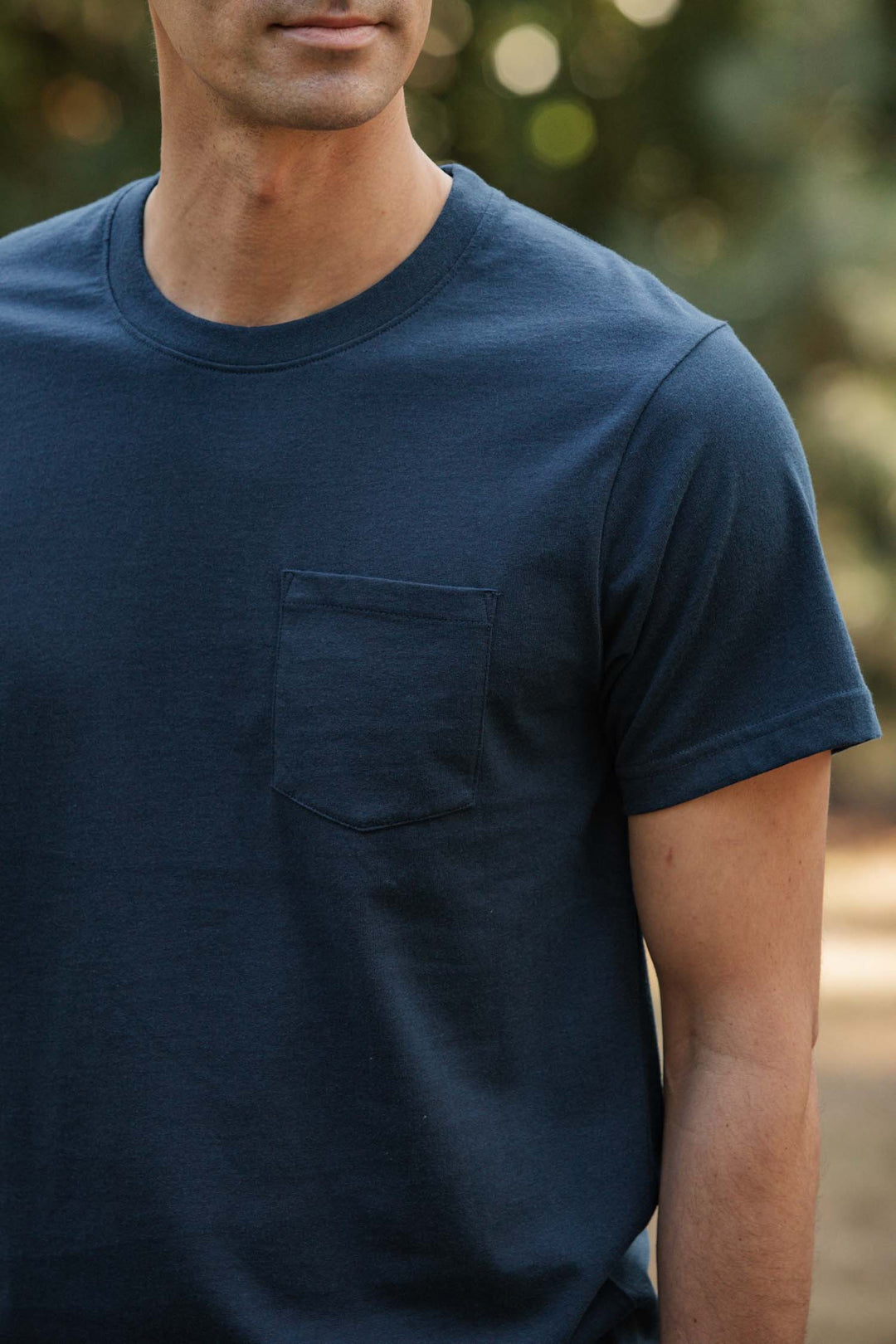 A man stands outdoors in a Bridge & Burn Recycled Pocket Tee in Cobalt, featuring a chest pocket; sunlight and blurred greenery create a vintage atmosphere.