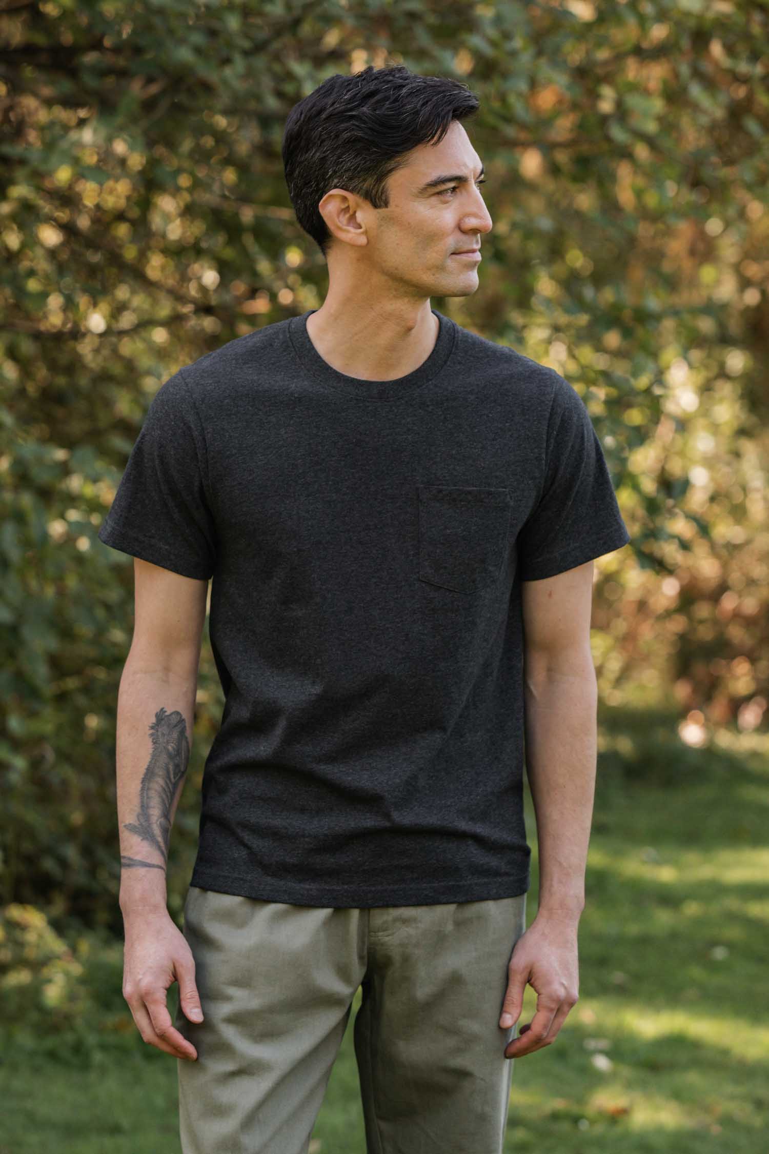 A man with short dark hair and a visible arm tattoo stands on grass with greenery behind him, wearing the Bridge & Burn Recycled Pocket Tee in Charcoal Heather and light pants, looking to the side.