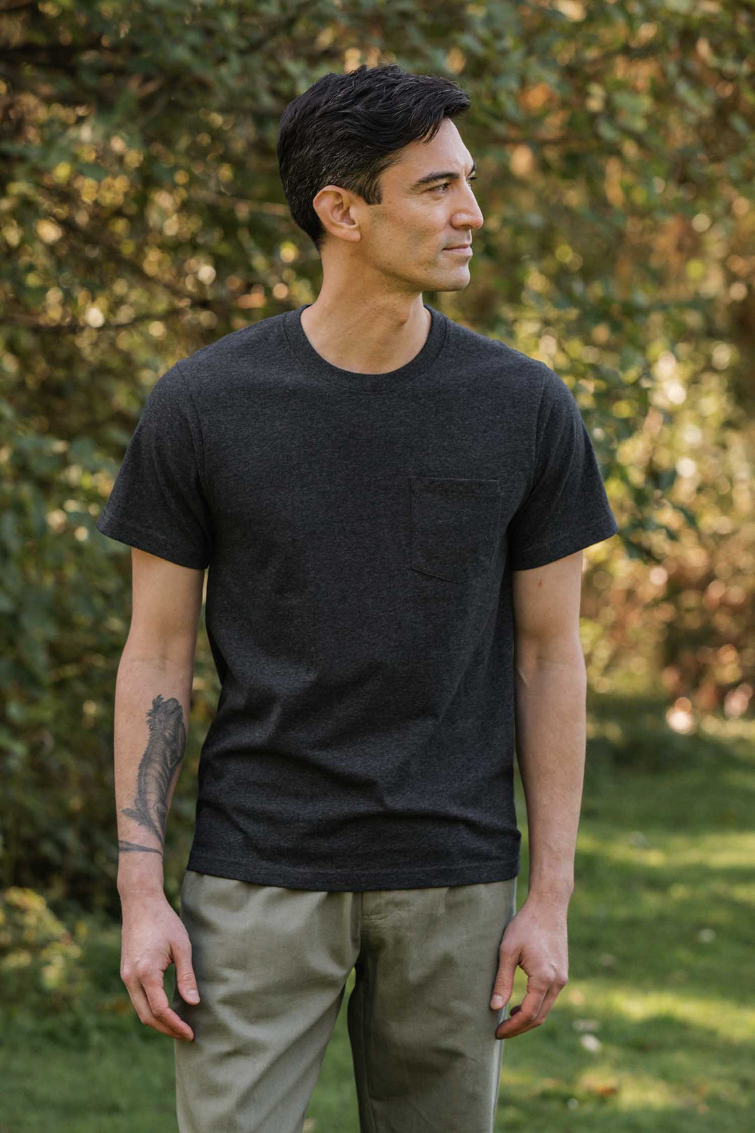 A man with short dark hair and a visible arm tattoo stands on grass with greenery behind him, wearing the Bridge & Burn Recycled Pocket Tee in Charcoal Heather and light pants, looking to the side.
